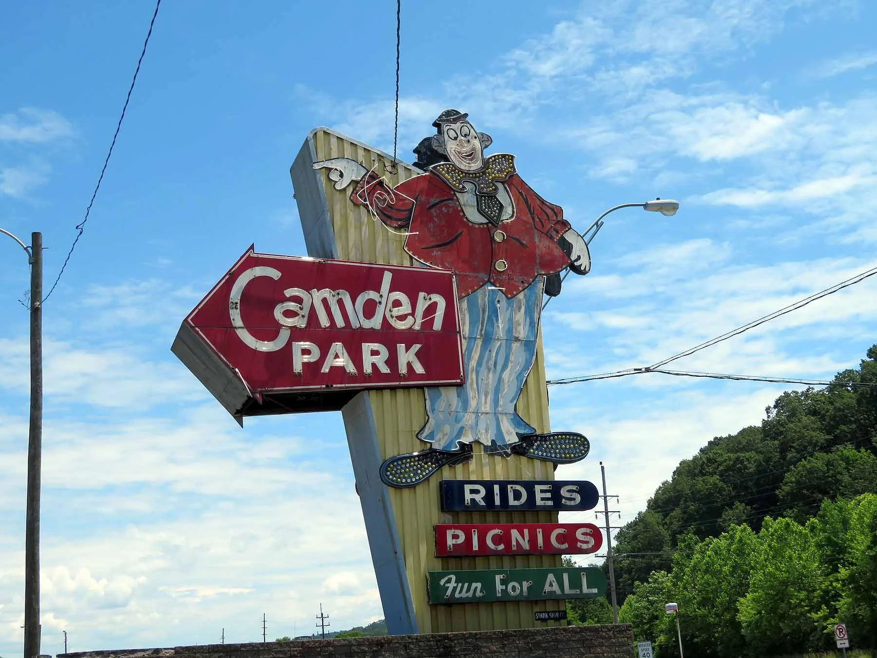 Vintage neon sign for Camden Park amusement park featuring a clown in a red jacket and striped pants, with signage indicating rides, picnics, and fun for all, against a partly cloudy sky with green trees in the background.