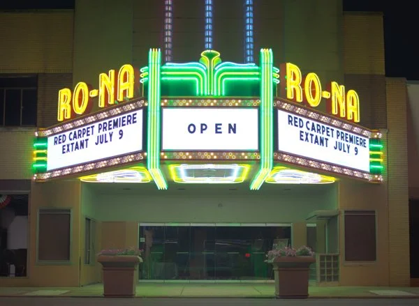 Neon-lit marquee theater sign for 'Ro-Na' with a digital display announcing 'Open' and information about a new movie premiering July 9.