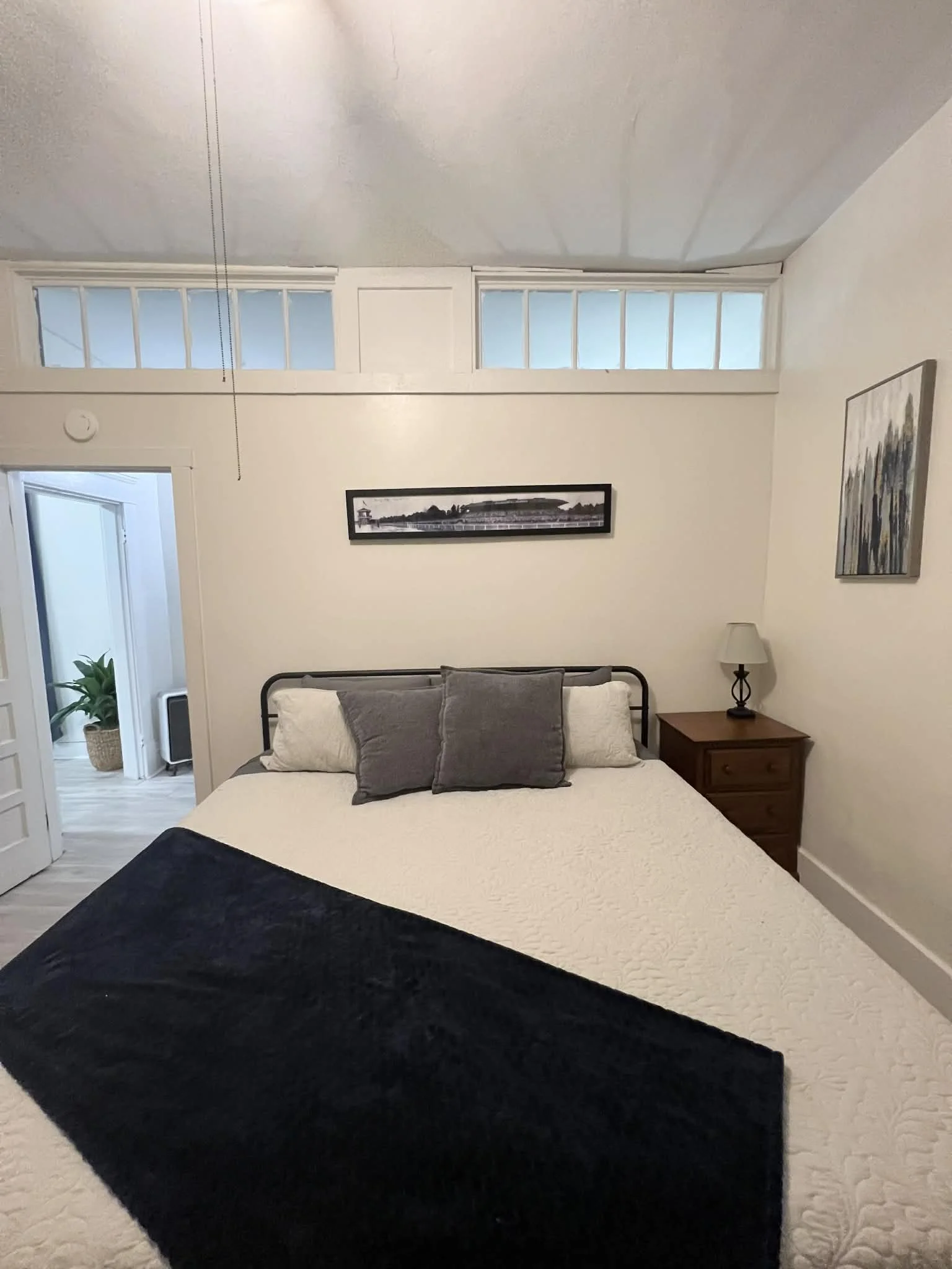 A bedroom with a bed featuring white bedding and three pillows, one white and two gray. There is a wooden nightstand with a lamp on it to the right of the bed. Art prints hang on the walls, and high windows above the wall let in natural light. An open door shows a room with a plant in a woven basket.