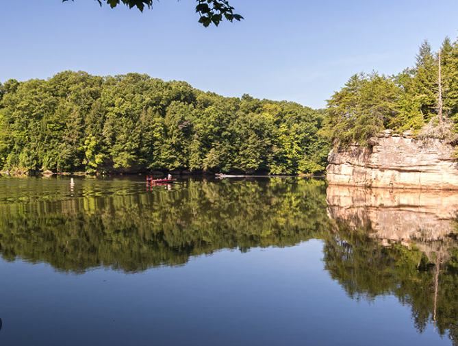 A calm Lake Vesuvius with dense green trees on the opposite bank and a rocky cliff on the right side, reflecting in the water under a clear blue sky.