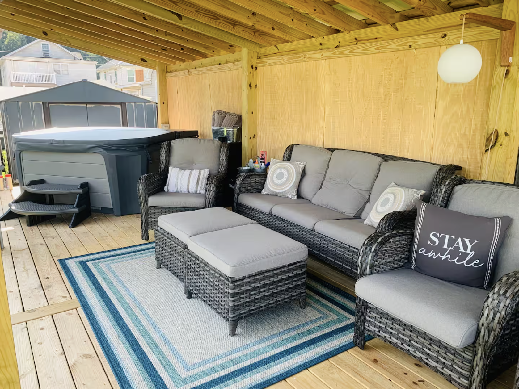Outdoor patio area with wicker sofa, armchair, ottoman, decorative pillows, a hot tub, wooden walls and ceiling, a hanging light, and a colorful striped area rug.