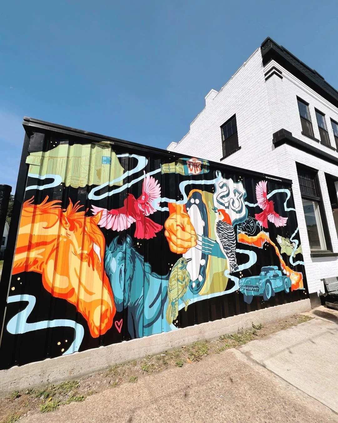 Colorful mural on a black metal building wall shows birds flying among horses, a vintage Corvette, and abstract shapes against a clear blue sky.