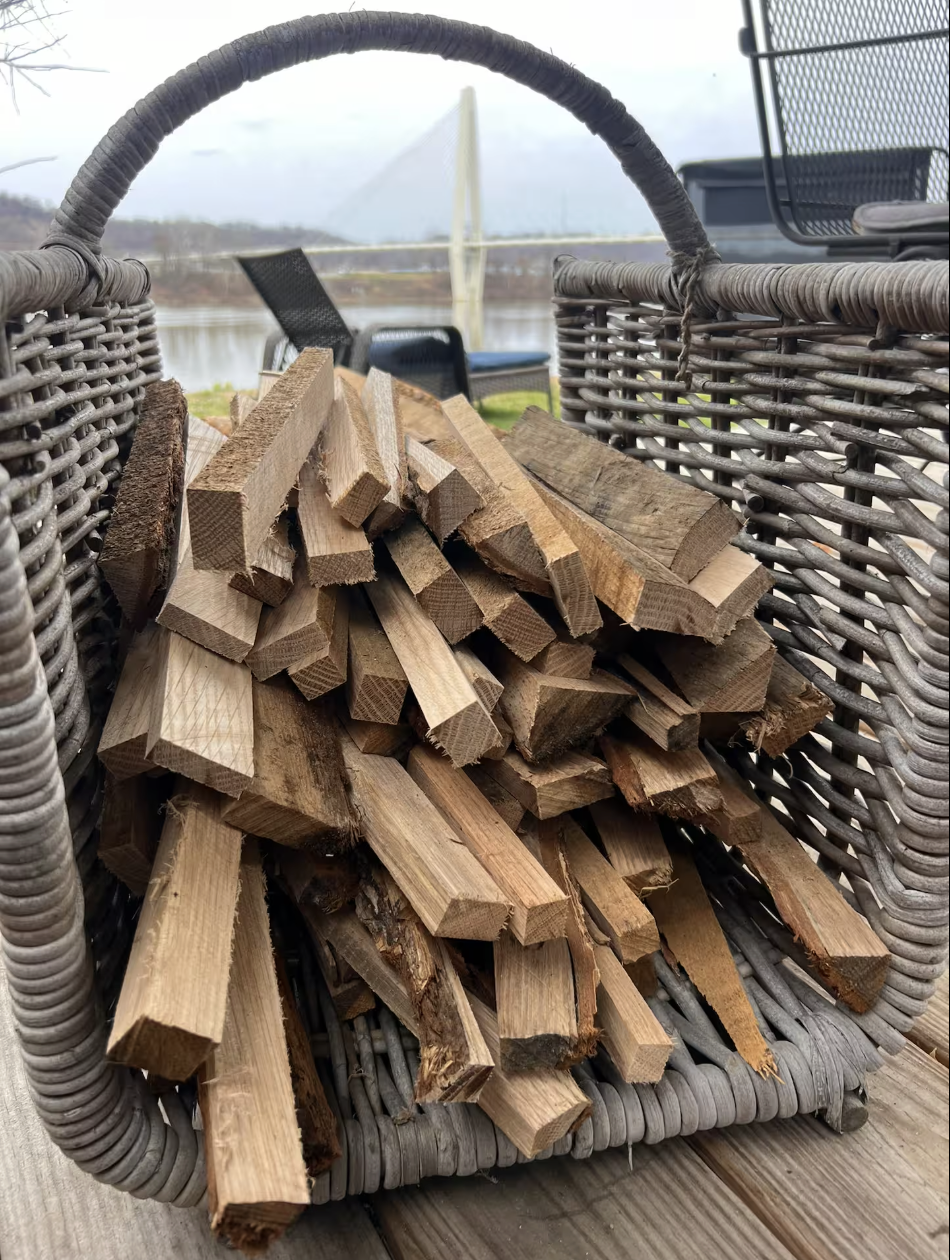 A wicker basket filled with chopped firewood resting on a wooden surface outdoors near a river, with chairs and a bridge visible in the background.