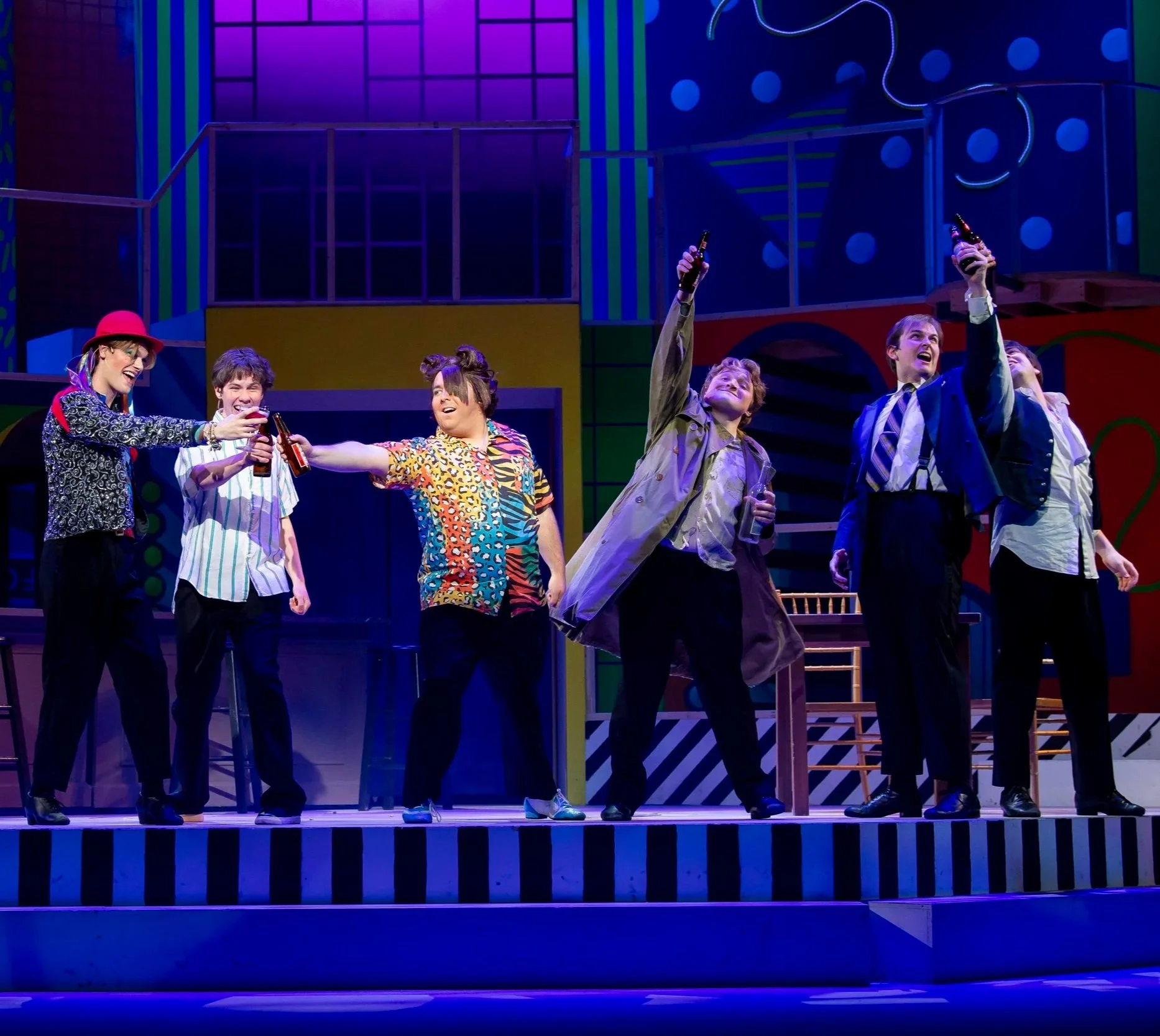 Actors on stage in a colorful set, holding beer bottles, celebrating, and raising their glasses in a toast during a theatrical performance.