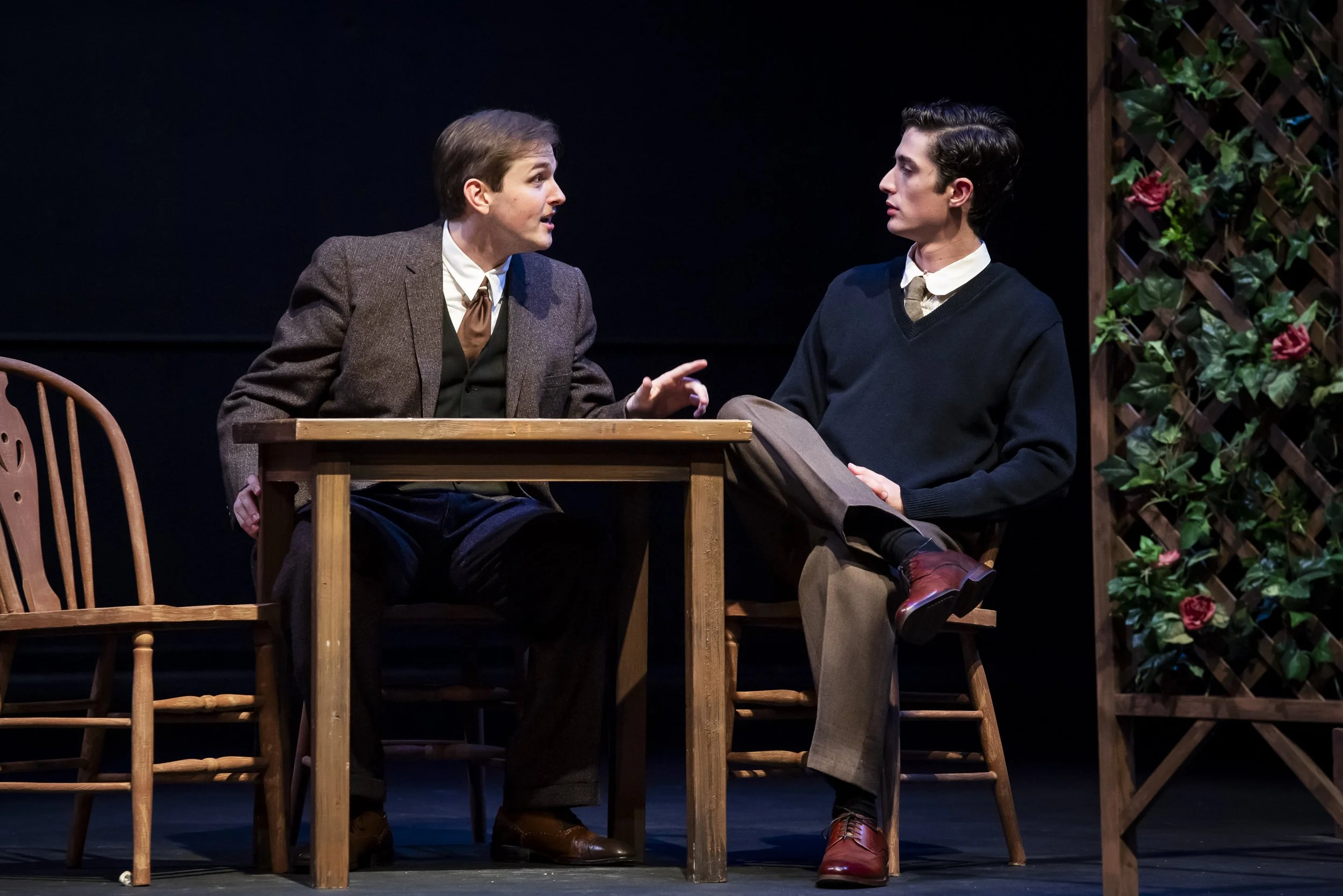 Two men in vintage-style clothing engaged in conversation at a wooden table on stage, with one gesturing and the other listening thoughtfully, in a theatrical setting with a dark backdrop and a trellis with pink roses on the side.