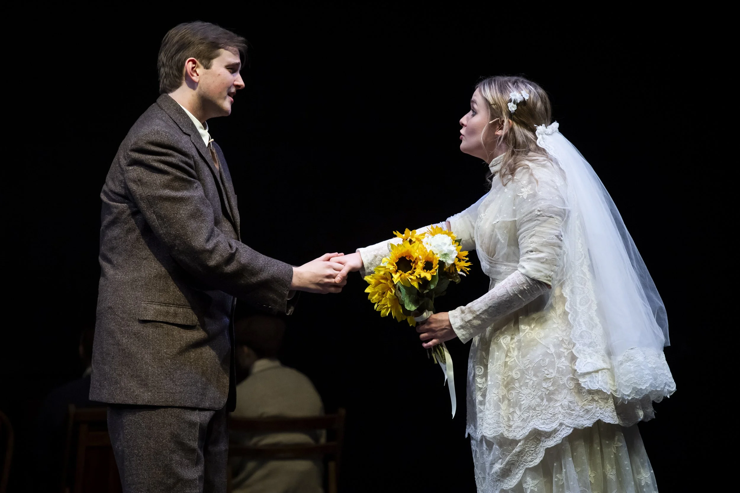 A man and woman in wedding attire exchanging vows or a handshake on stage, with the woman holding a bouquet of sunflowers, against a black background.