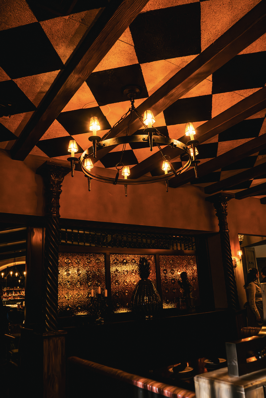Interior of a dimly lit restaurant with a circular chandelier with lit bulbs hanging from a checkered ceiling with orange and black squares. Decorative columns with twisted designs frame a backlit wall with floral or patterned glass, and some people are visible in the background.