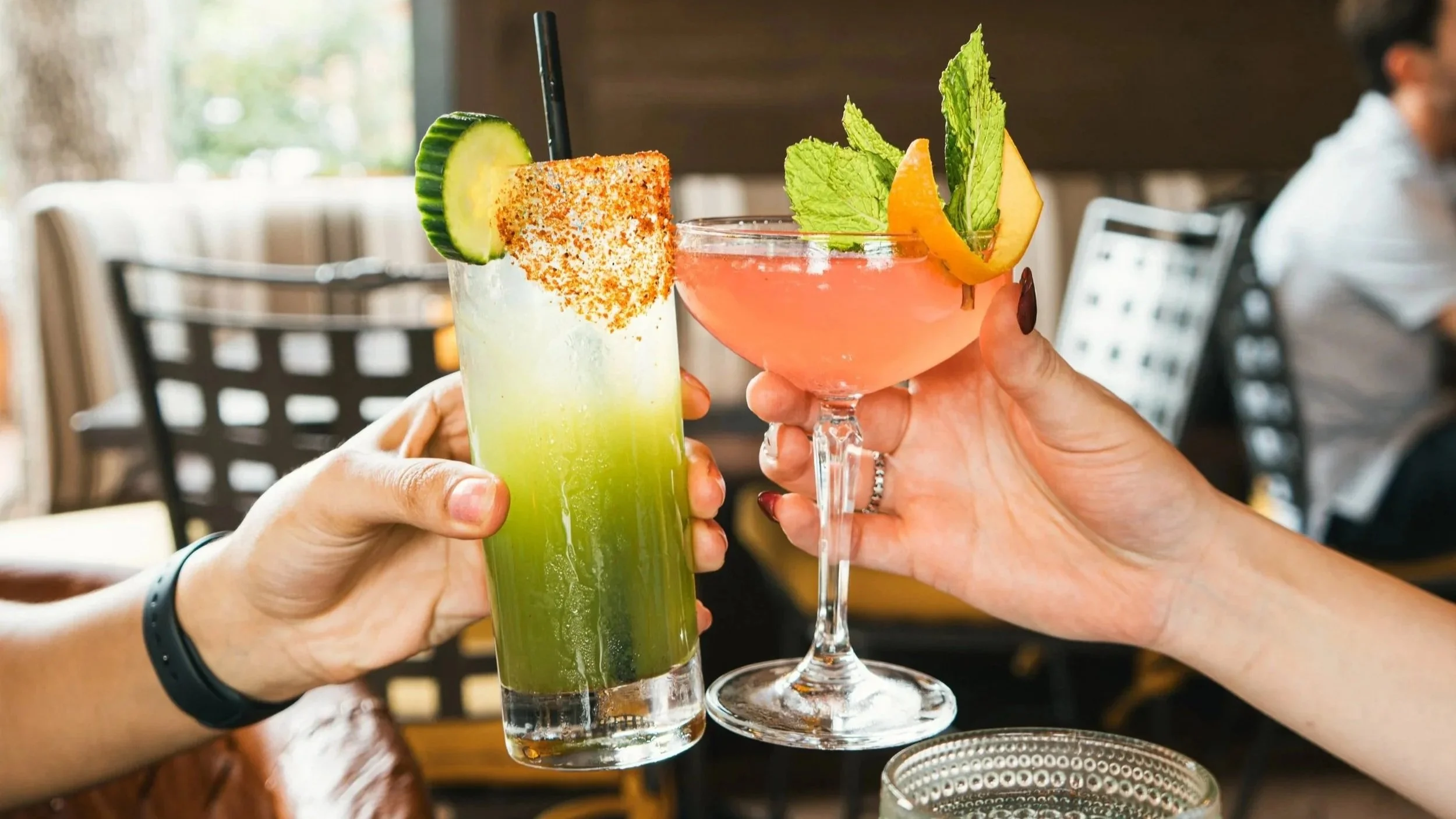 Two people clinking mimosas with garnishes, one with a green cocktail topped with cucumber and chili powder, and the other with a pink cocktail garnished with mint leaves and an orange slice, in a restaurant.