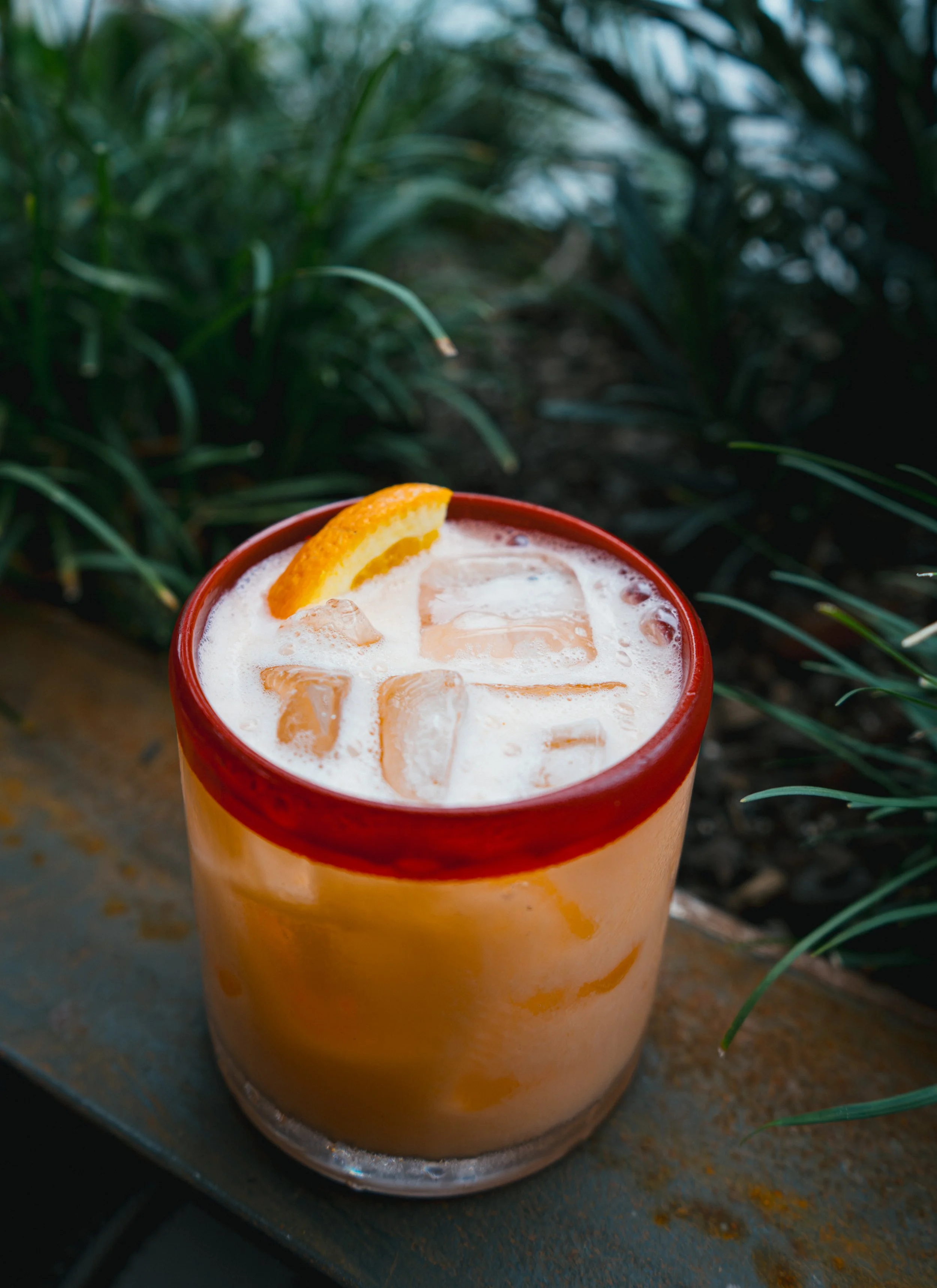 A glass of iced orange beverage with a slice of orange on top, set on a rustic surface with green foliage in the background.