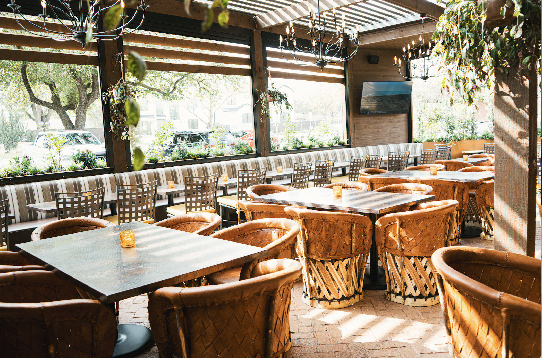 Sunlit indoor restaurant dining area with wooden tables, leather chairs, large windows, and decorative chandeliers.