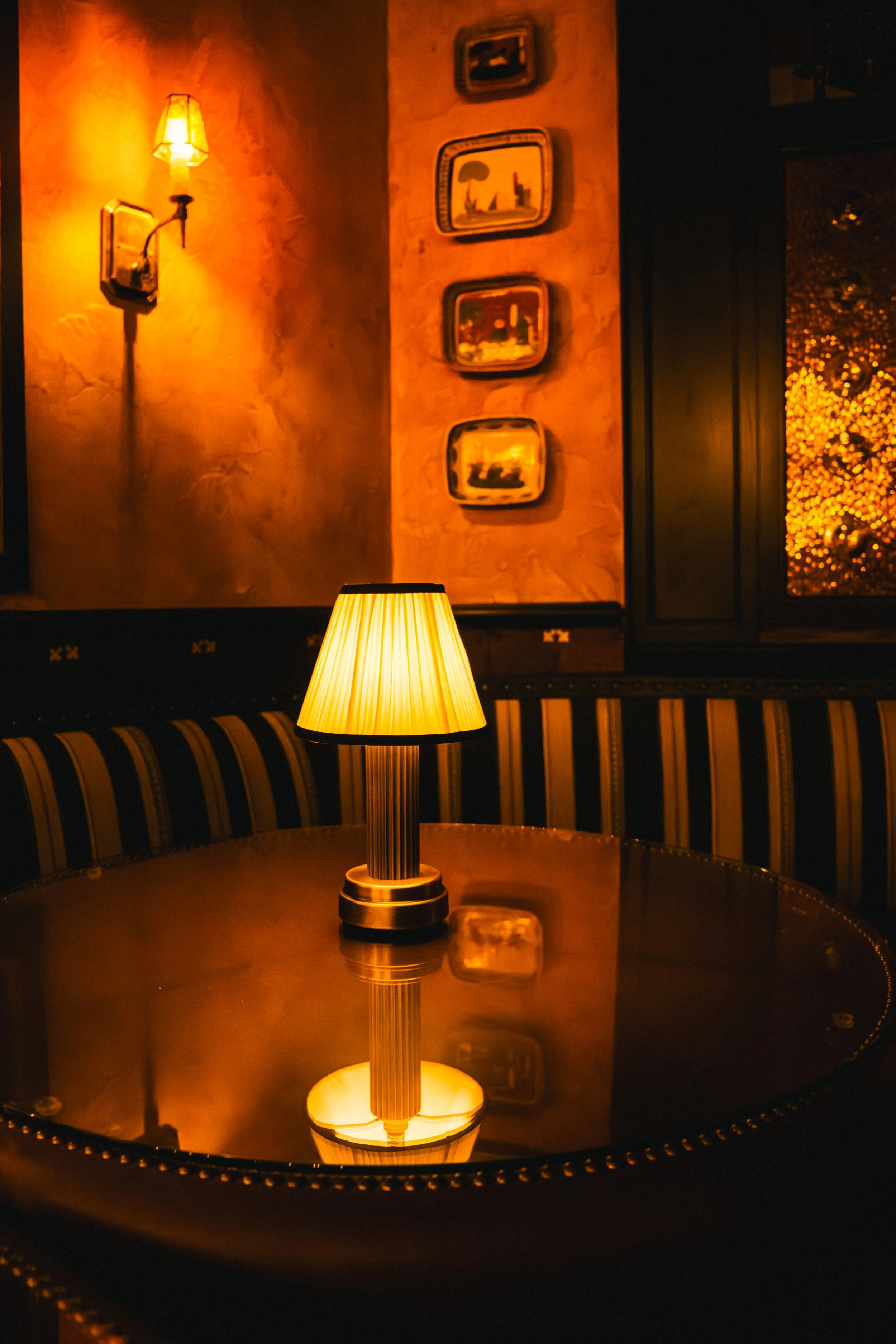 A warm, dimly lit room with a table lamp on a polished wooden table, reflecting the lamp's light, and a dark striped upholstered booth. The wall features framed artwork and a wall-mounted light fixture.