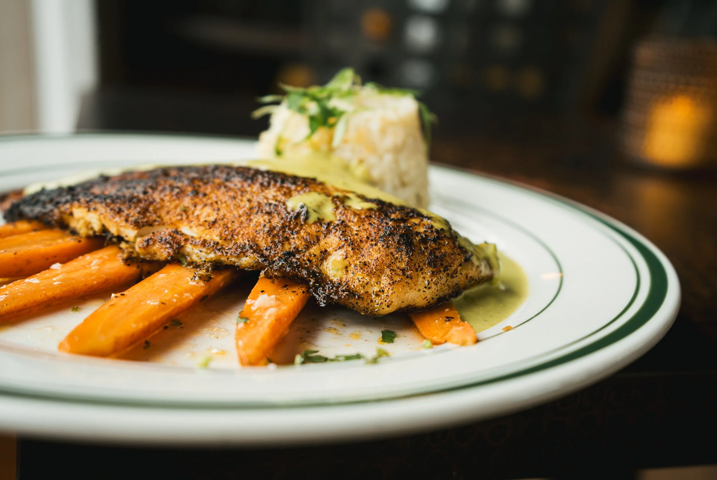 Blackened fish fillet served over roasted carrots with a side of mashed potatoes garnished with herbs.
