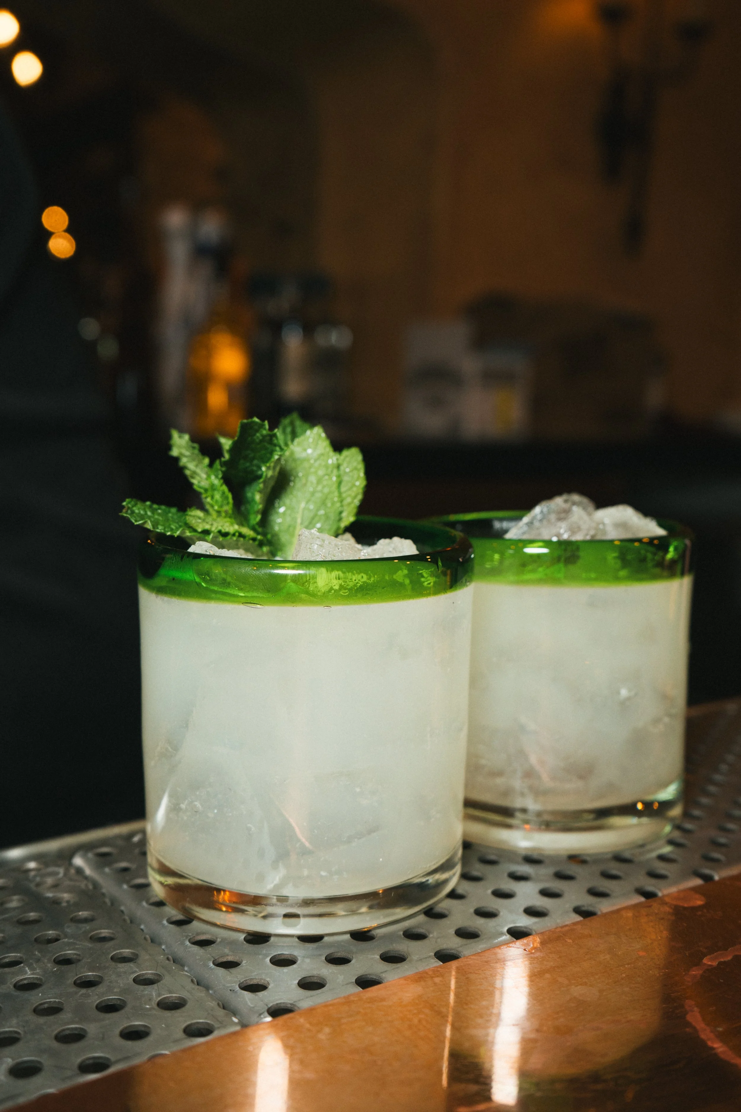 Two cocktails with mint garnish on a bar counter, featuring clear glasses with ice and green rims.