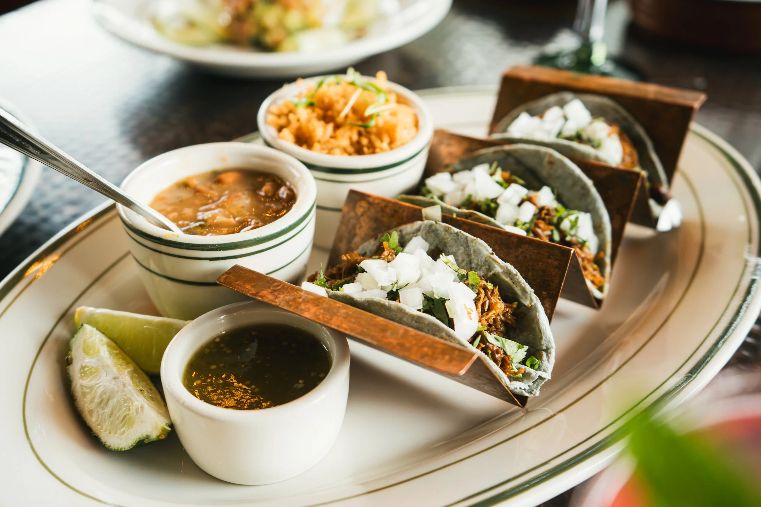 Three tacos with shredded meat, chopped onions, and herbs on a white platter, accompanied by lime wedges, a cup of salsa verde, and two small bowls of beans and rice.