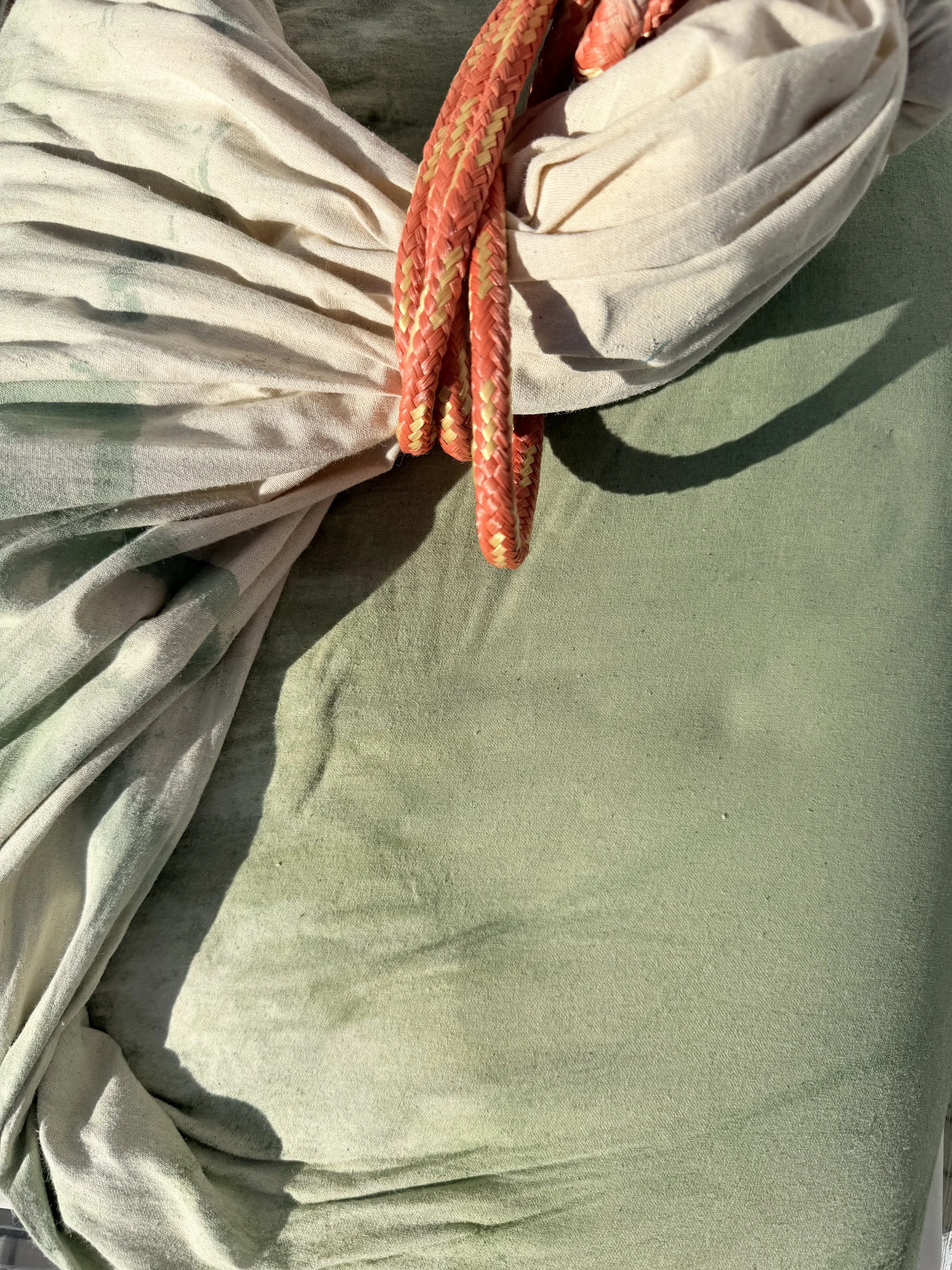 Close-up of a person's arm with a light-colored long-sleeve shirt, secured with a pink and yellow rope. The background is a light green fabric surface.