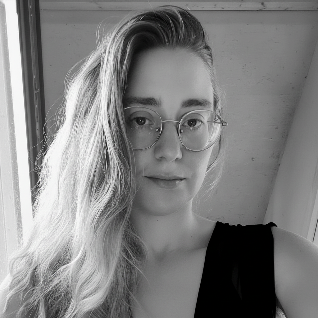 A young woman with long wavy hair and glasses taking a selfie in front of a window