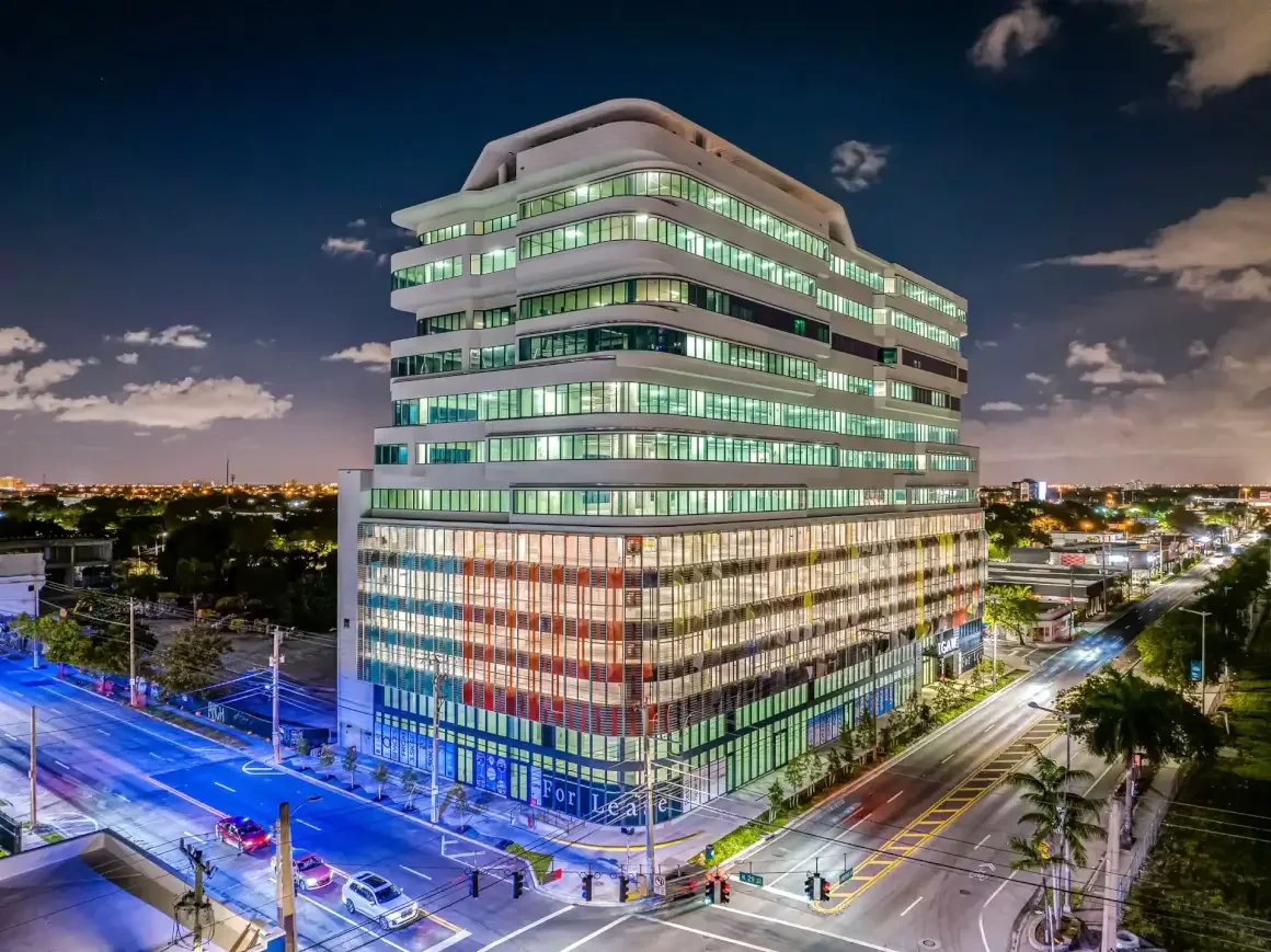 Night view of a modern multi-story office building with illuminated windows, situated on a city street with cars and palm trees.