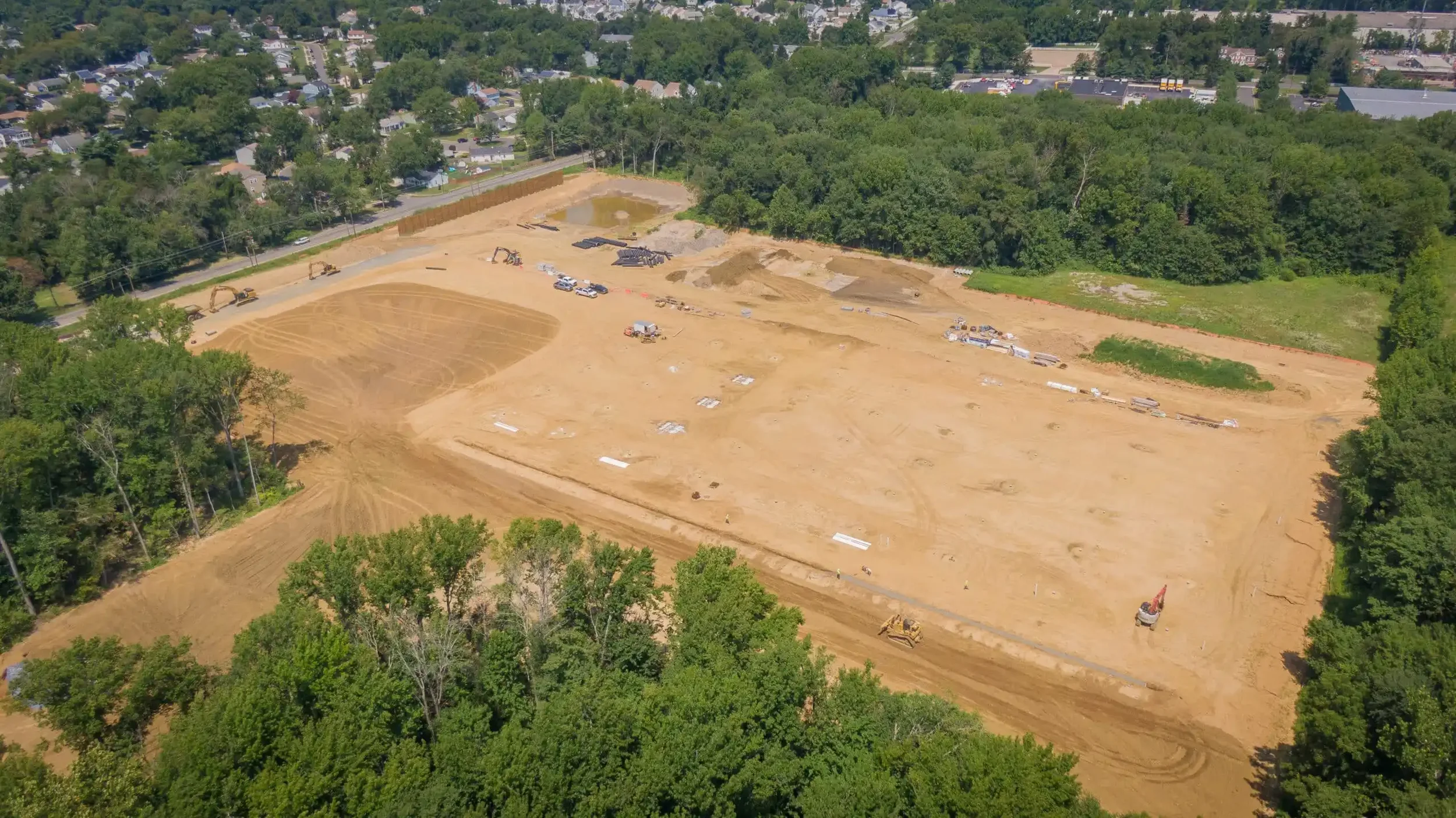 An aerial view of a construction site with cleared land, construction vehicles, and equipment, surrounded by trees and nearby residential and commercial areas.
