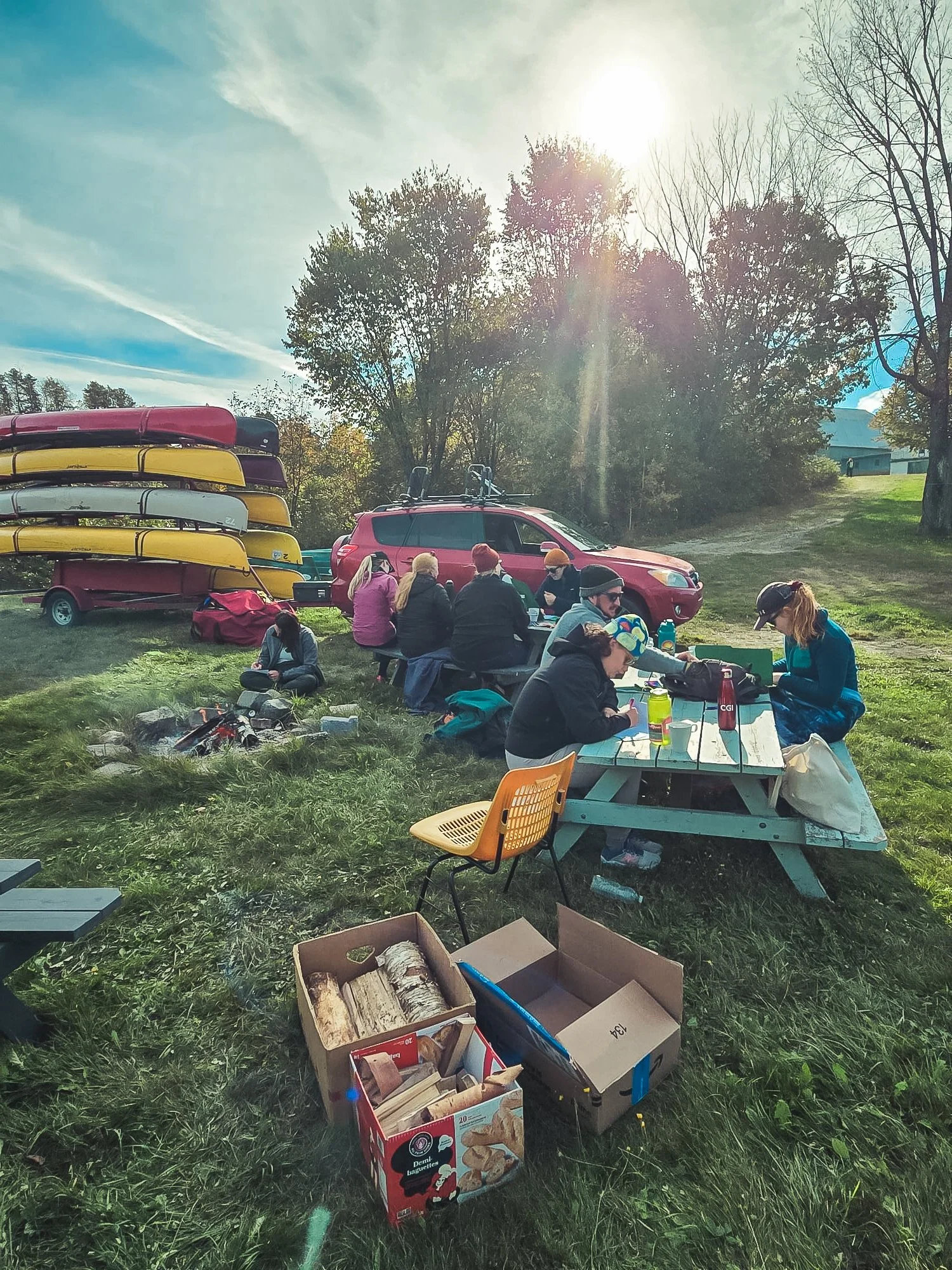 Groupe de personnes assises autour d'une table en plein air, avec des kayaks empilés sur un chariot et une voiture sur l'herbe, soleil brillant dans le ciel.