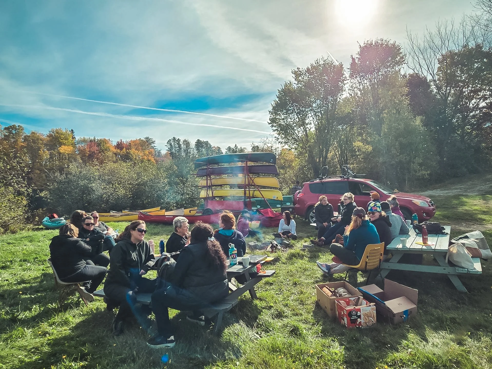 Groupe de personnes assises en cercle lors d'un pique-nique en plein air avec des kayaks empilés en arrière-plan et un véhicule rouge sous un ciel ensoleillé.