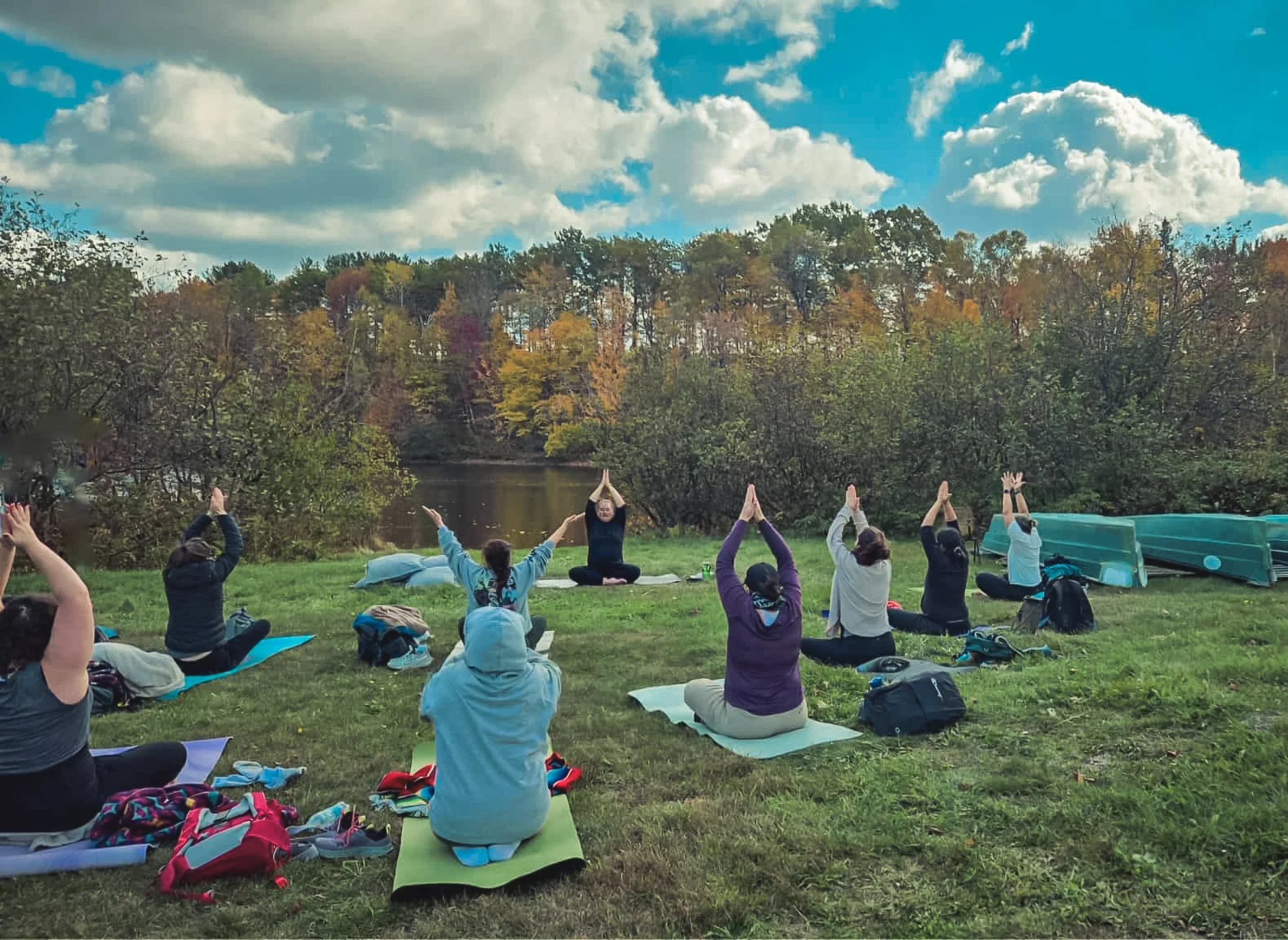 Groupe de personnes pratiquant le yoga en plein air sur l'herbe près d'une rivière, sous un ciel partiellement nuageux, avec des arbres aux couleurs d'automne en arrière-plan.