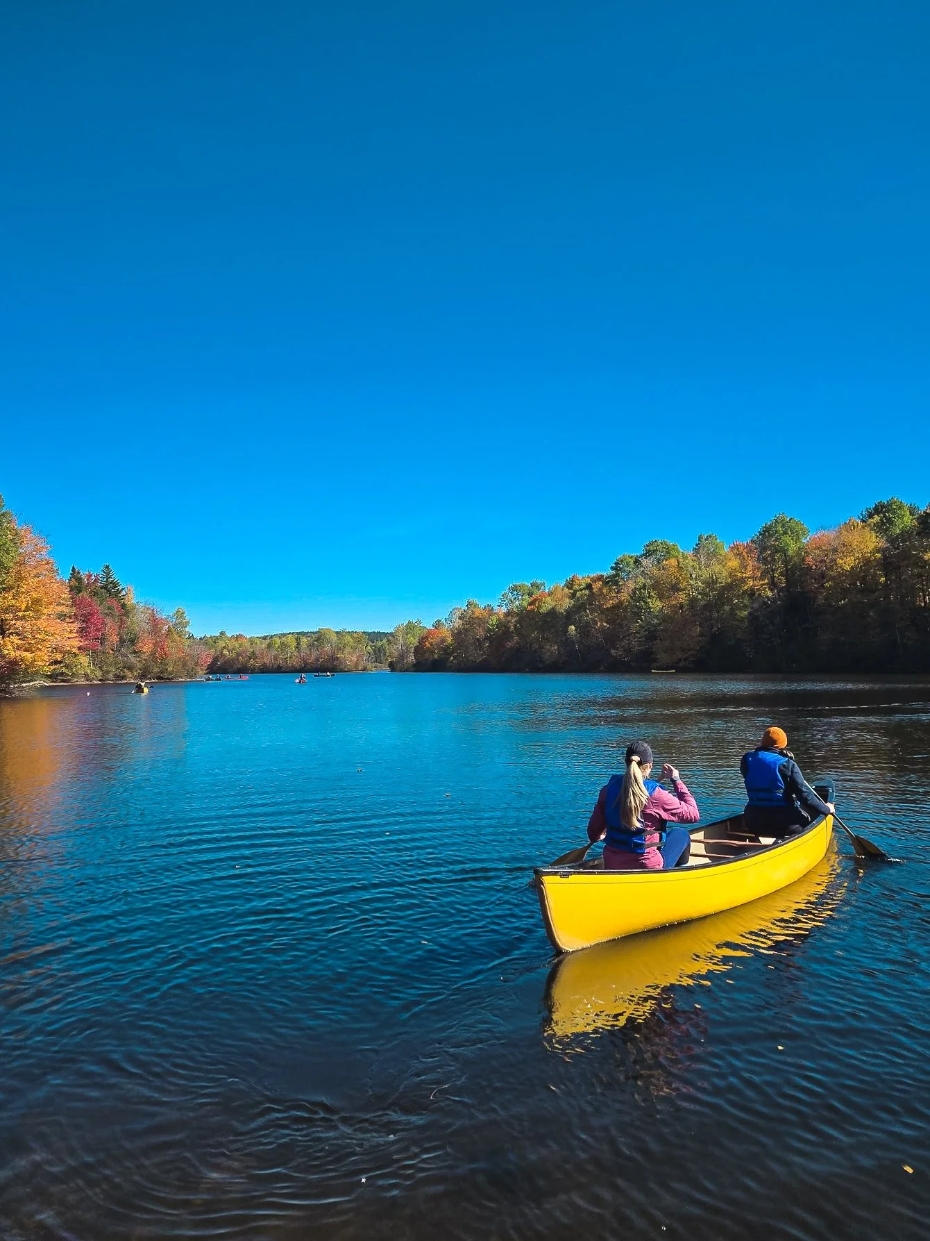 Deux personnes en kayak jaune naviguant sur un lac avec des arbres aux couleurs d'automne sous un ciel bleu clair.