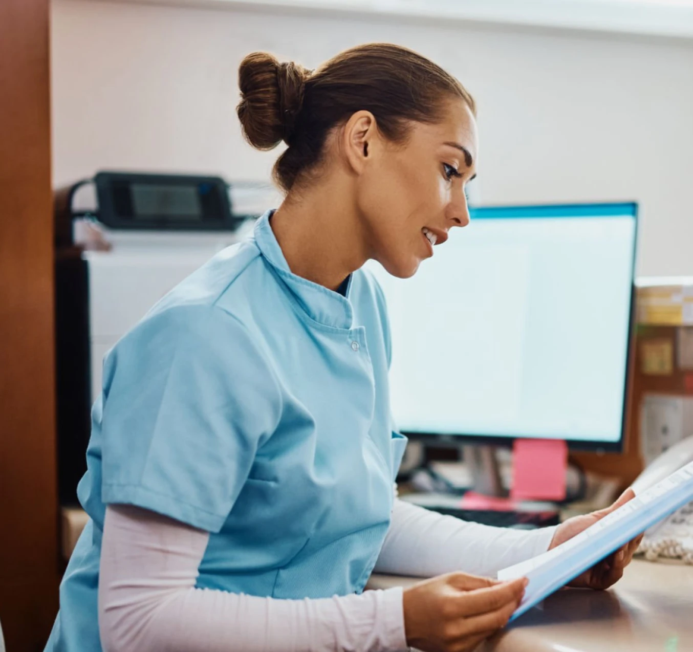 A female healthcare worker in light blue scrubs sitting at a desk, reading a document with a computer monitor and office supplies in the background.