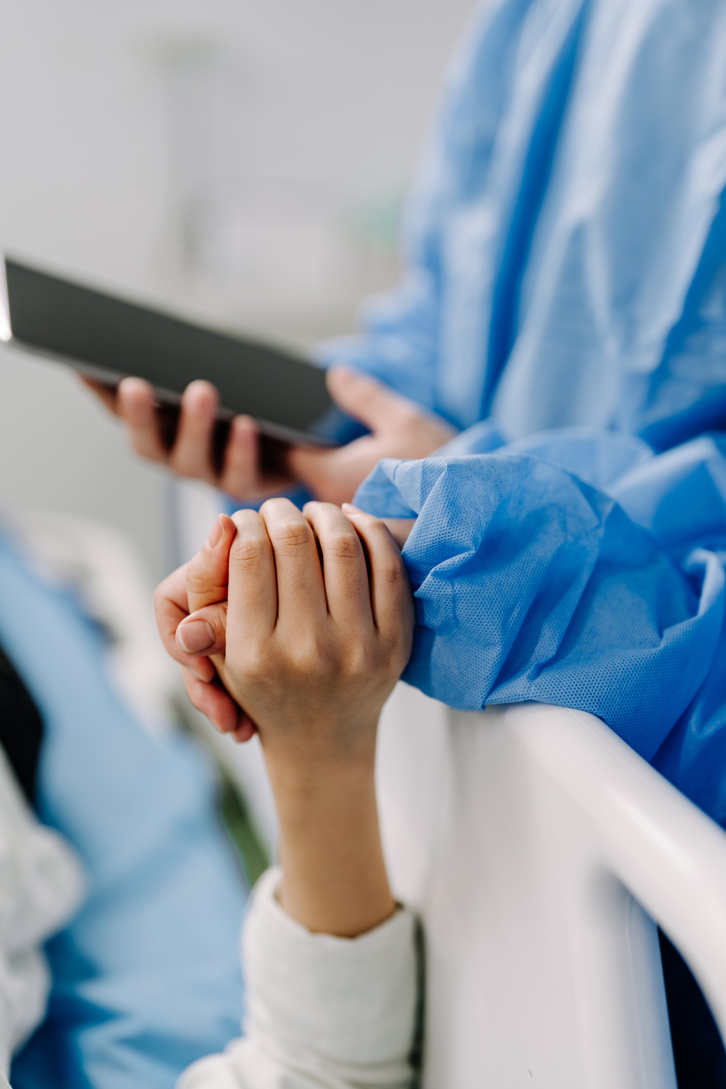 Close-up of two hands holding each other, one wearing a white sleeve, the other in blue medical scrubs. A healthcare worker in blue scrubs is visible in the background, using a tablet.