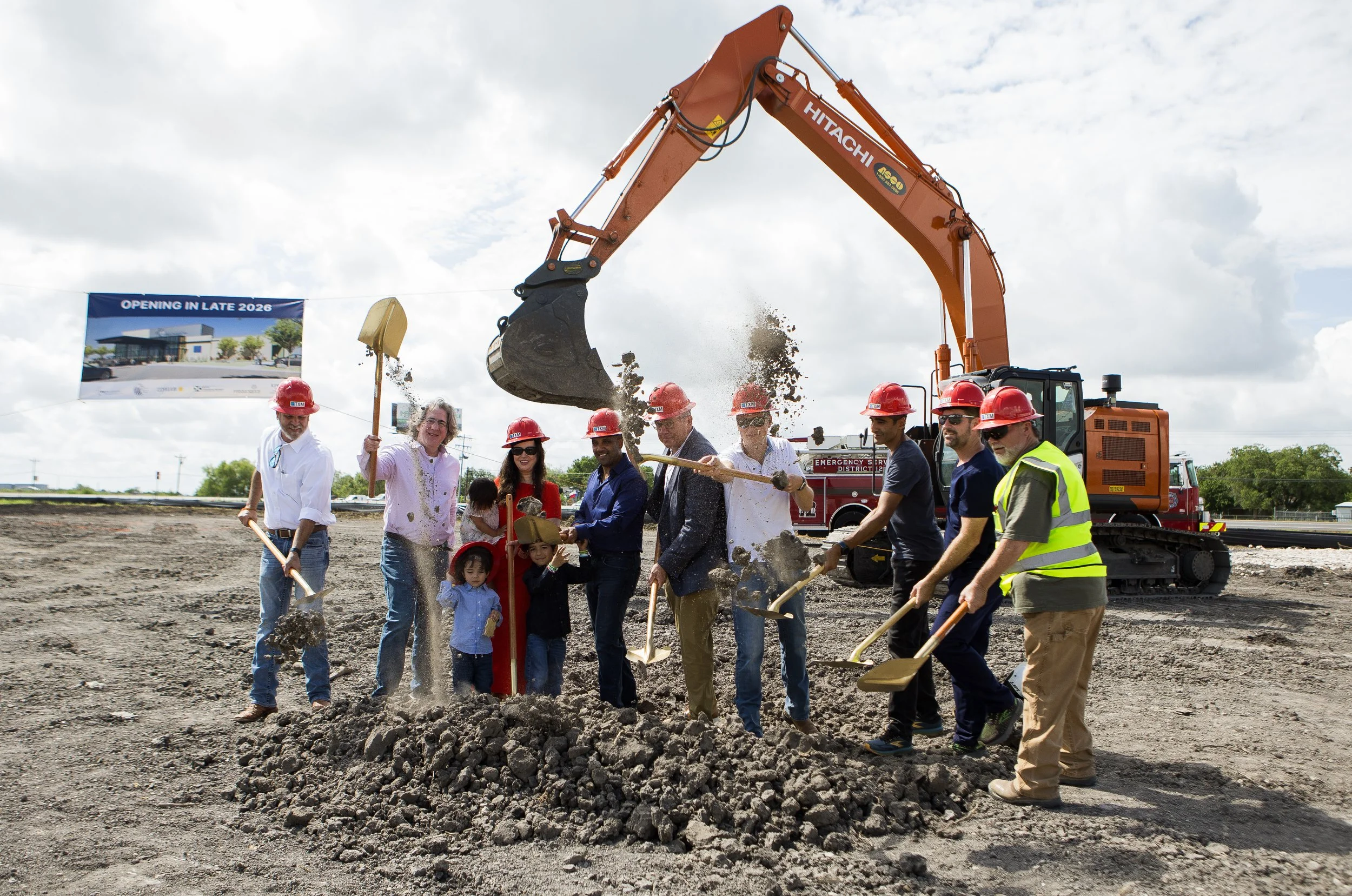 People participating in a groundbreaking ceremony for a new construction project, wearing red safety helmets, with a large excavator in the background and a banner that says 'Opening in Late 2026'.