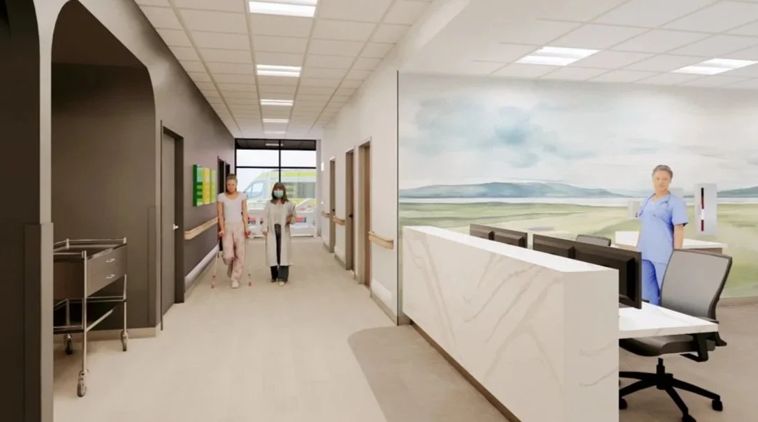 Hospital corridor with two women, one in pink scrubs and one in a gray coat, walking with crutches; a nurse in blue scrubs standing behind a reception desk near a large landscape mural.