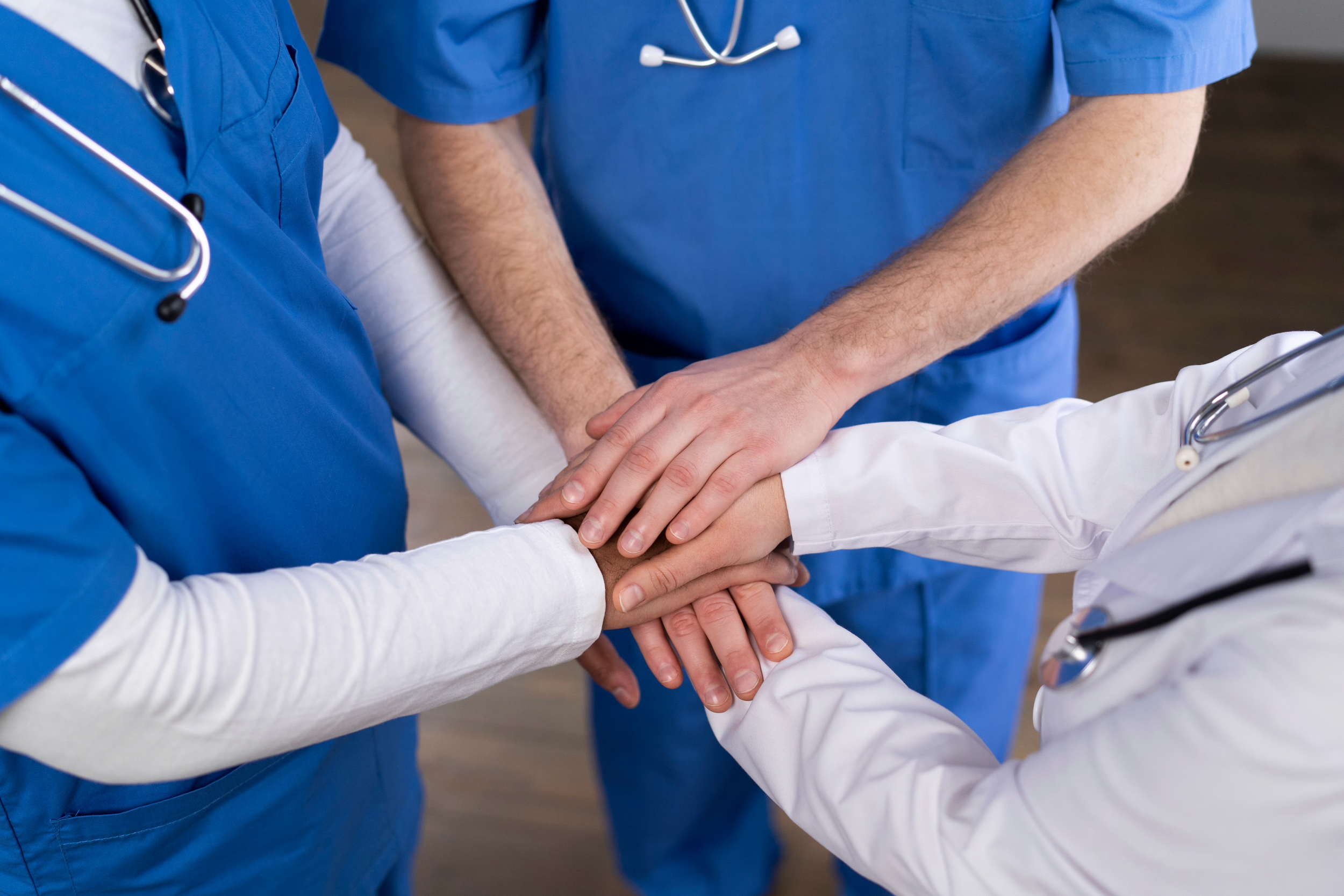 Medical team in blue scrubs and white coat placing their hands together in a huddle.