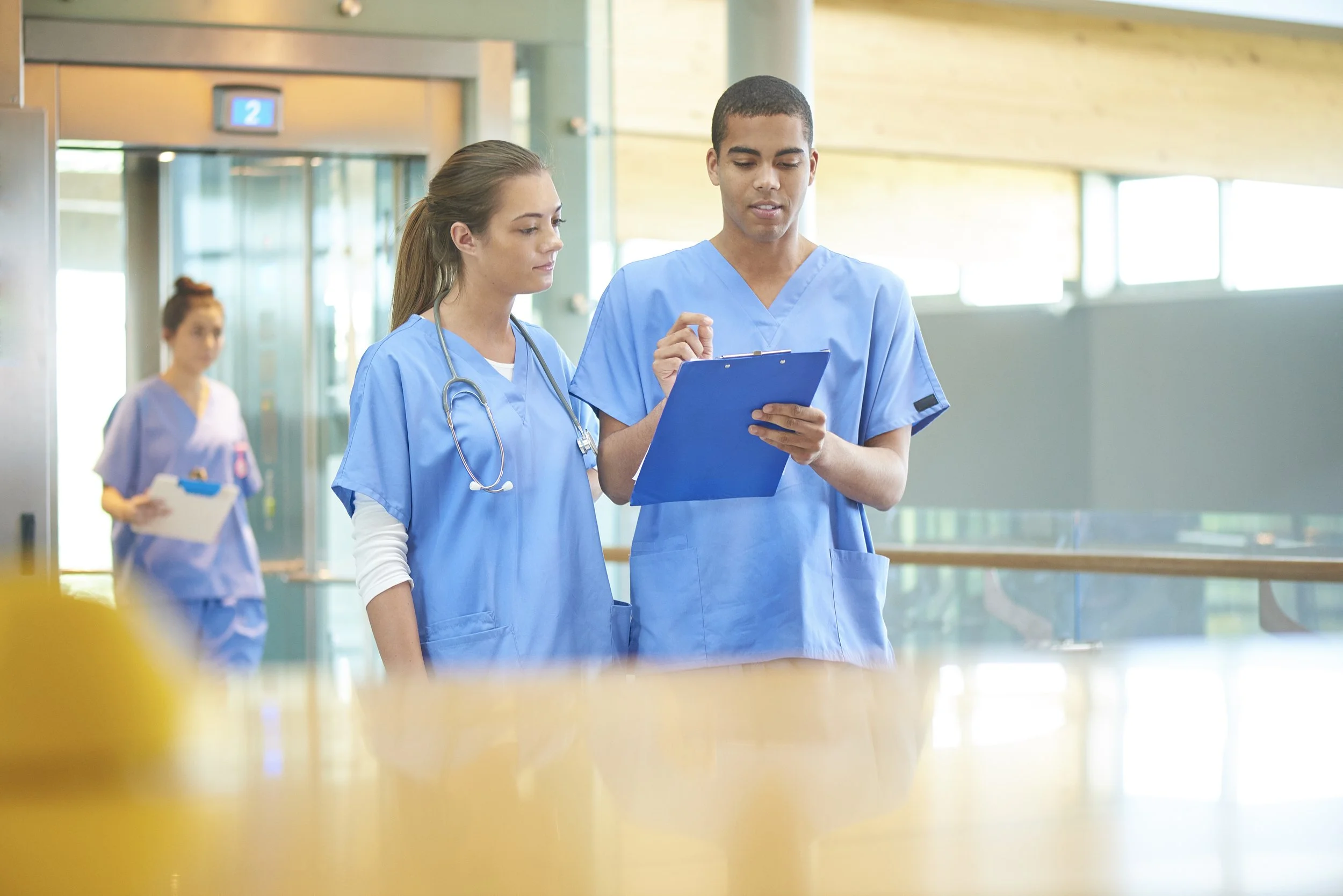 Two healthcare professionals, a woman and a man, in blue scrubs reviewing documents on a clipboard in a hospital corridor, with another nurse in the background.