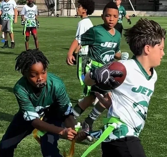 Children playing youth football on a grassy field, wearing green and white jerseys with 'Jets' logo, with one child running with a football and another reaching for the ball.
