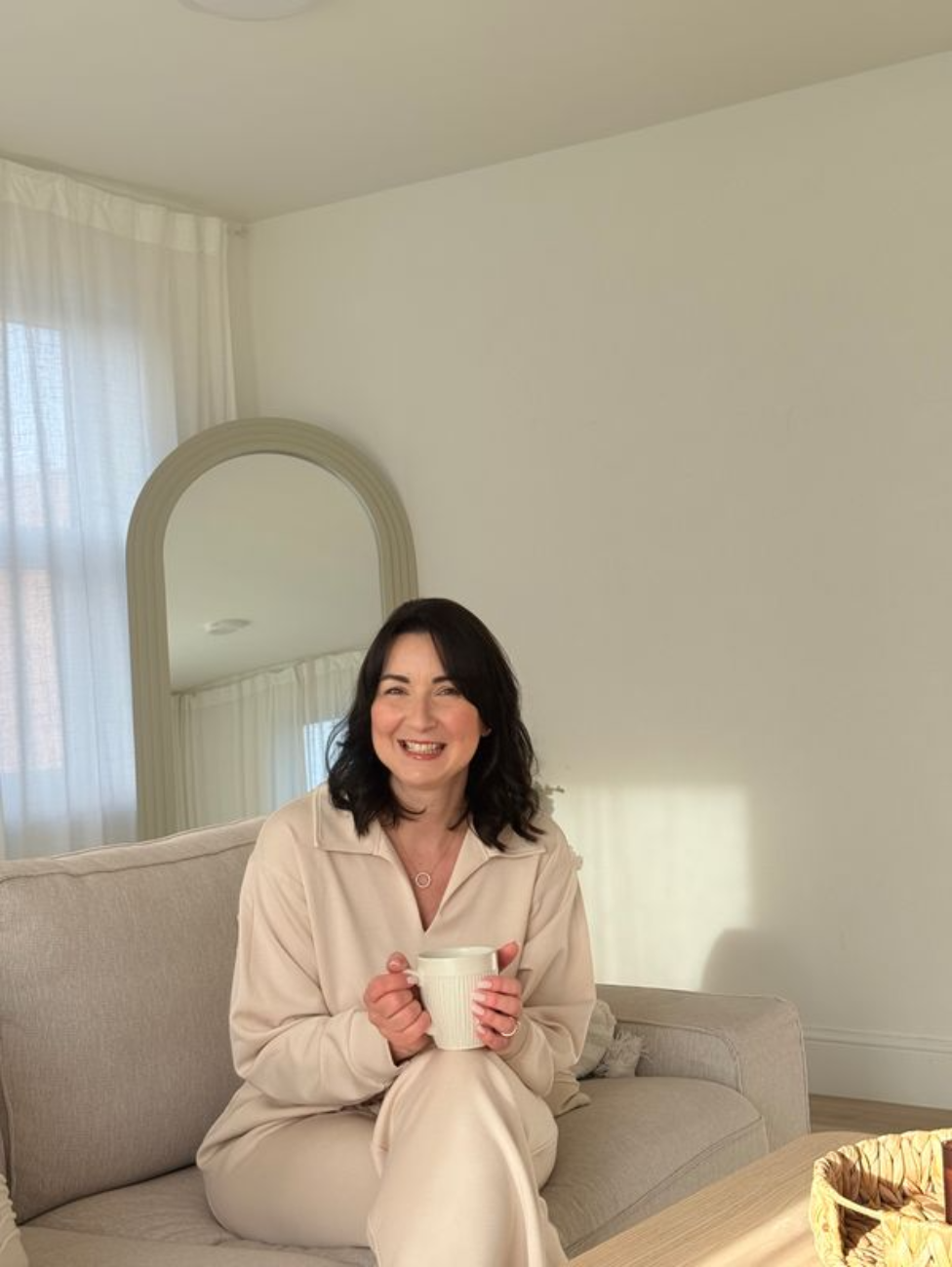 A woman with black hair smiling and holding a white mug, sitting on a beige couch in a bright room with white walls and curtains.