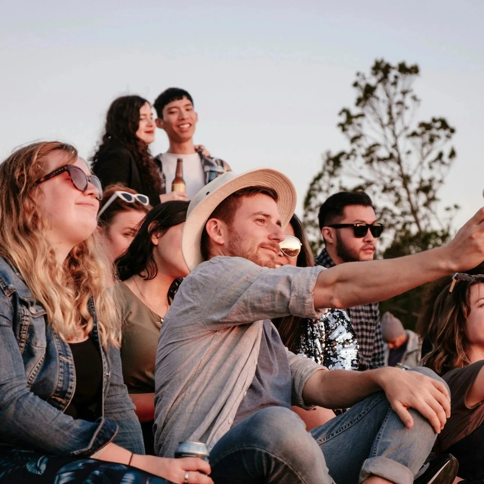 Group of friends enjoying an outdoor concert during sunset, taking selfies and posing with drinks and snacks.