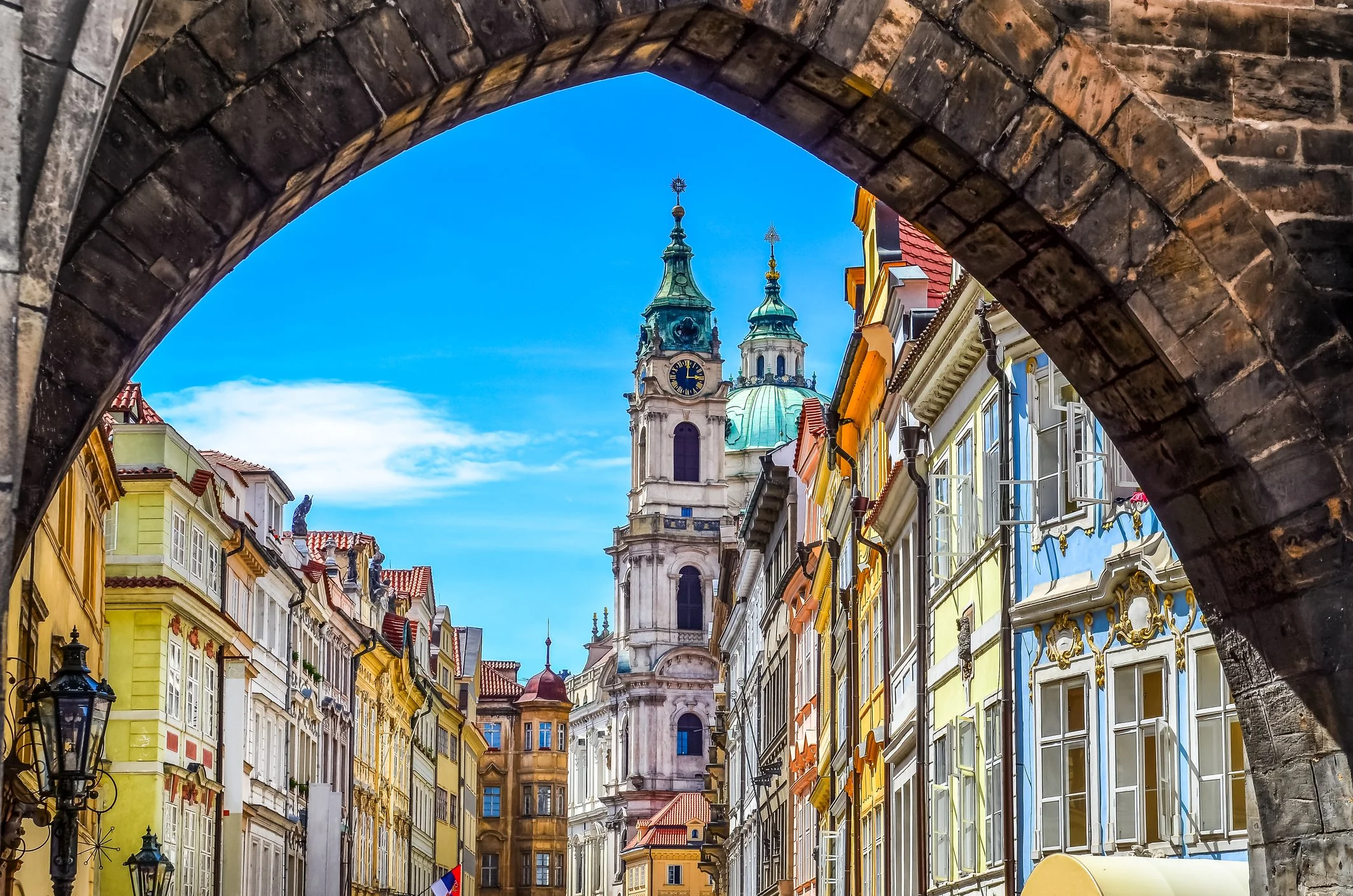 View of a historic European city street through an archway, with colorful buildings and a church tower with clock and domes in the background, under a clear blue sky.