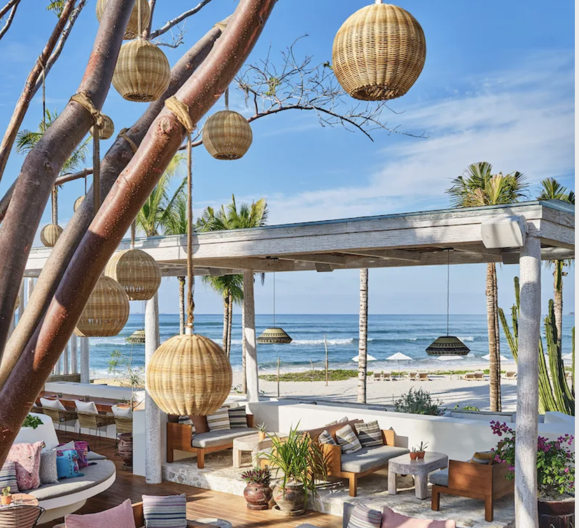 Beachside outdoor lounge area with hanging wicker lamps, wooden furniture, colorful cushions, and a view of the ocean with palm trees and a blue sky.