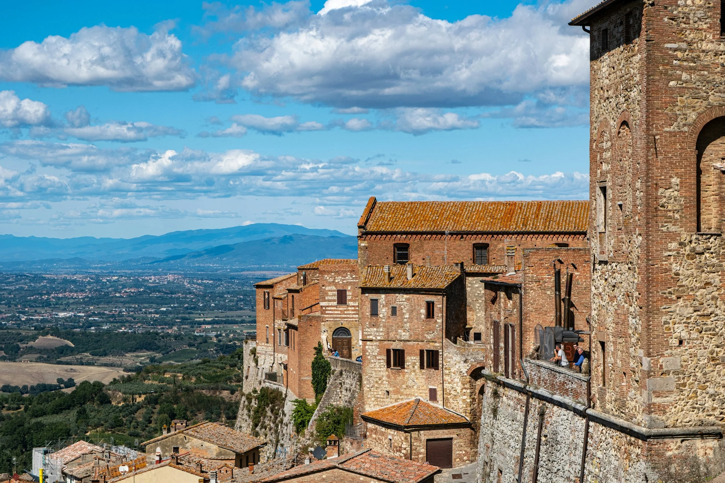 A scenic view of an ancient brick and stone castle or fortress perched on a hill, overlooking a vast landscape of fields and mountains under a partly cloudy sky. Three people are seen on a terrace of the castle enjoying the view.
