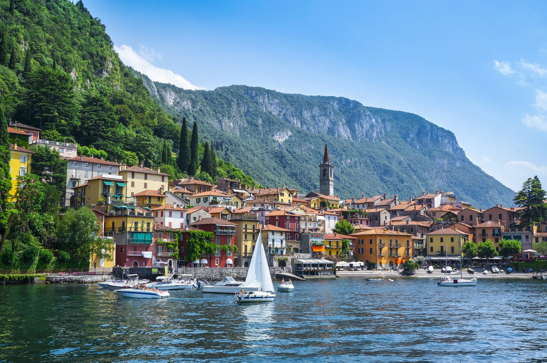 Colorful houses along a waterfront with boats, mountain in the background under a blue sky.