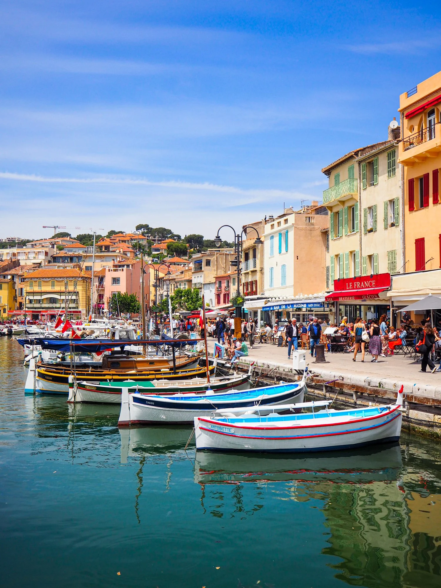 Colorful boats docked along a marina promenade with outdoor cafes and pastel-colored buildings in a lively seaside town under a clear blue sky.