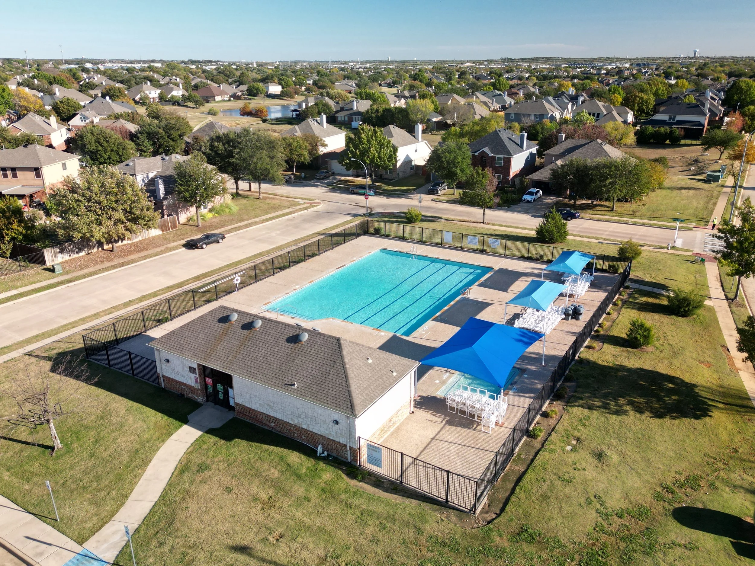 A community swimming pool with blue water, surrounded by a black fencing, with multiple blue shade canopies and white chairs arranged underneath, located in a residential neighborhood with houses and trees in the background under a clear sky.