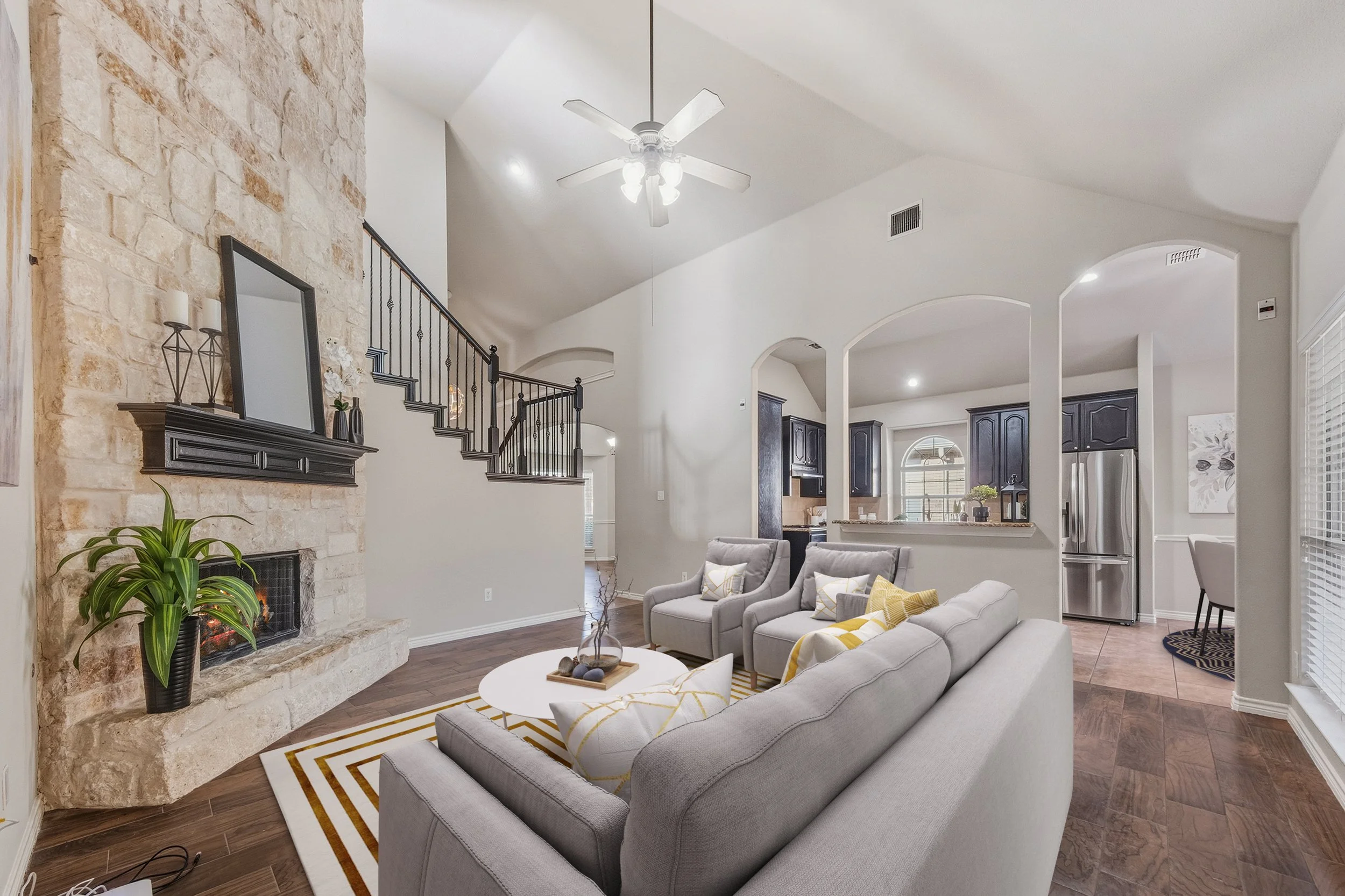 Living room with stone fireplace, light gray couch, two gray armchairs, coffee table, and kitchen in background with black cabinets and stainless steel refrigerator.