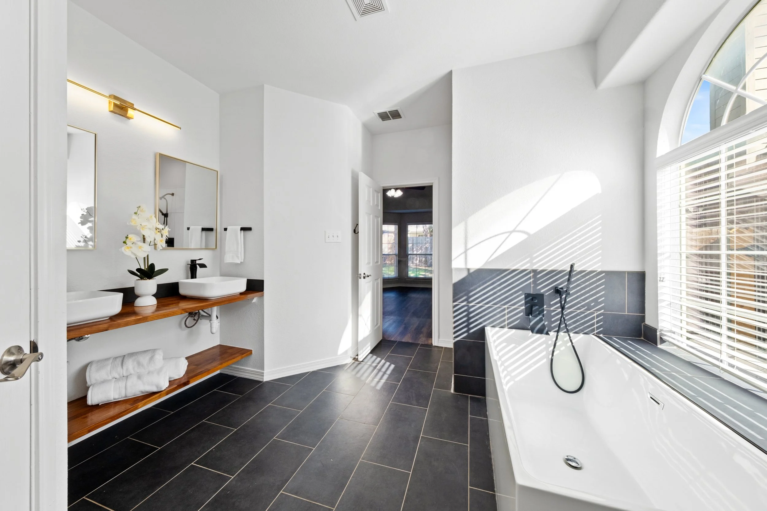 Bright bathroom with dark tile floor, double sinks on a wooden countertop, and a large bathtub under a window with blinds, featuring a modern, minimalist design.
