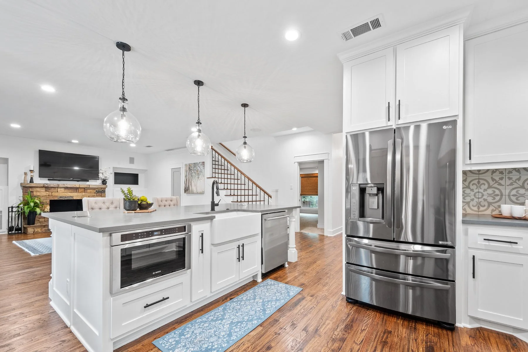 Modern kitchen with white cabinets, stainless steel refrigerator, island with sink, pendant lights, hardwood floors, and a view of the living room with a fireplace and TV.