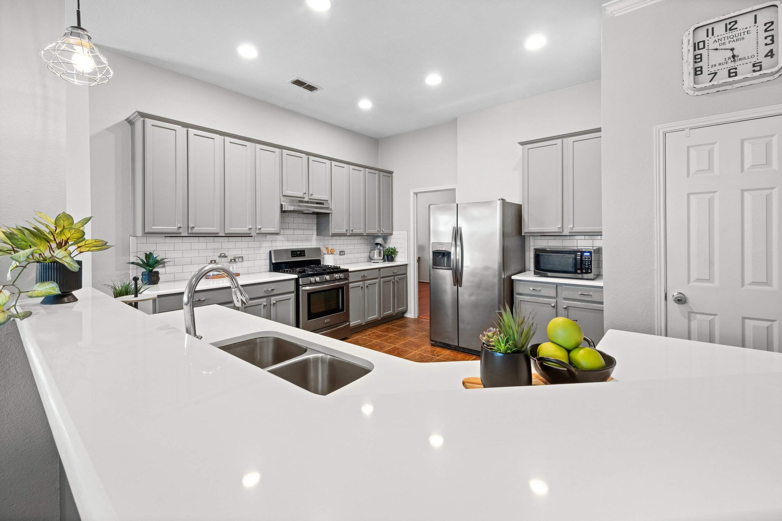 Modern kitchen with gray cabinets, stainless steel appliances, white subway tile backsplash, white countertop, and small potted plants. Includes refrigerator, microwave, stove, and sink area.