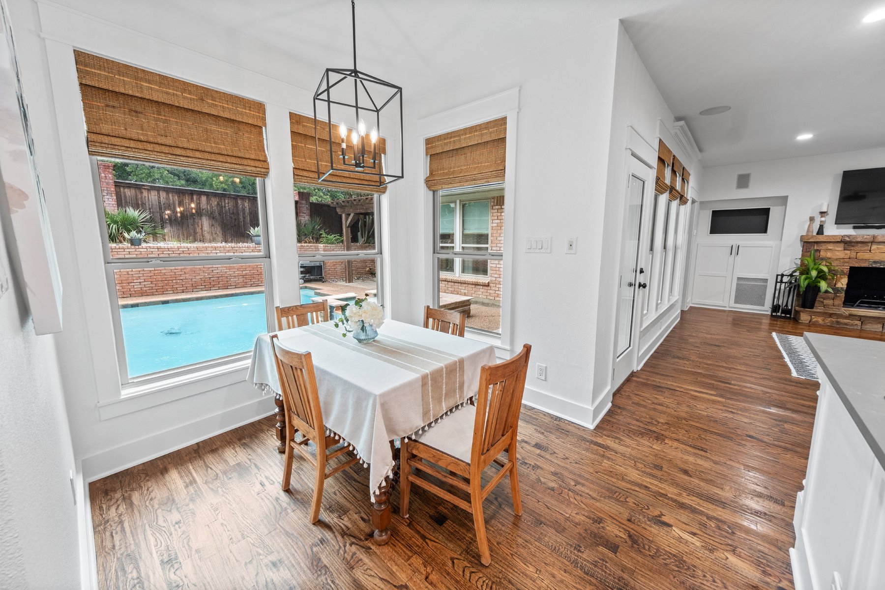 Dining area with a wooden table and four chairs, near large windows overlooking a swimming pool outside, with a brick wall in the background. The room has hardwood floors, white walls, and a modern black chandelier hanging from the ceiling.