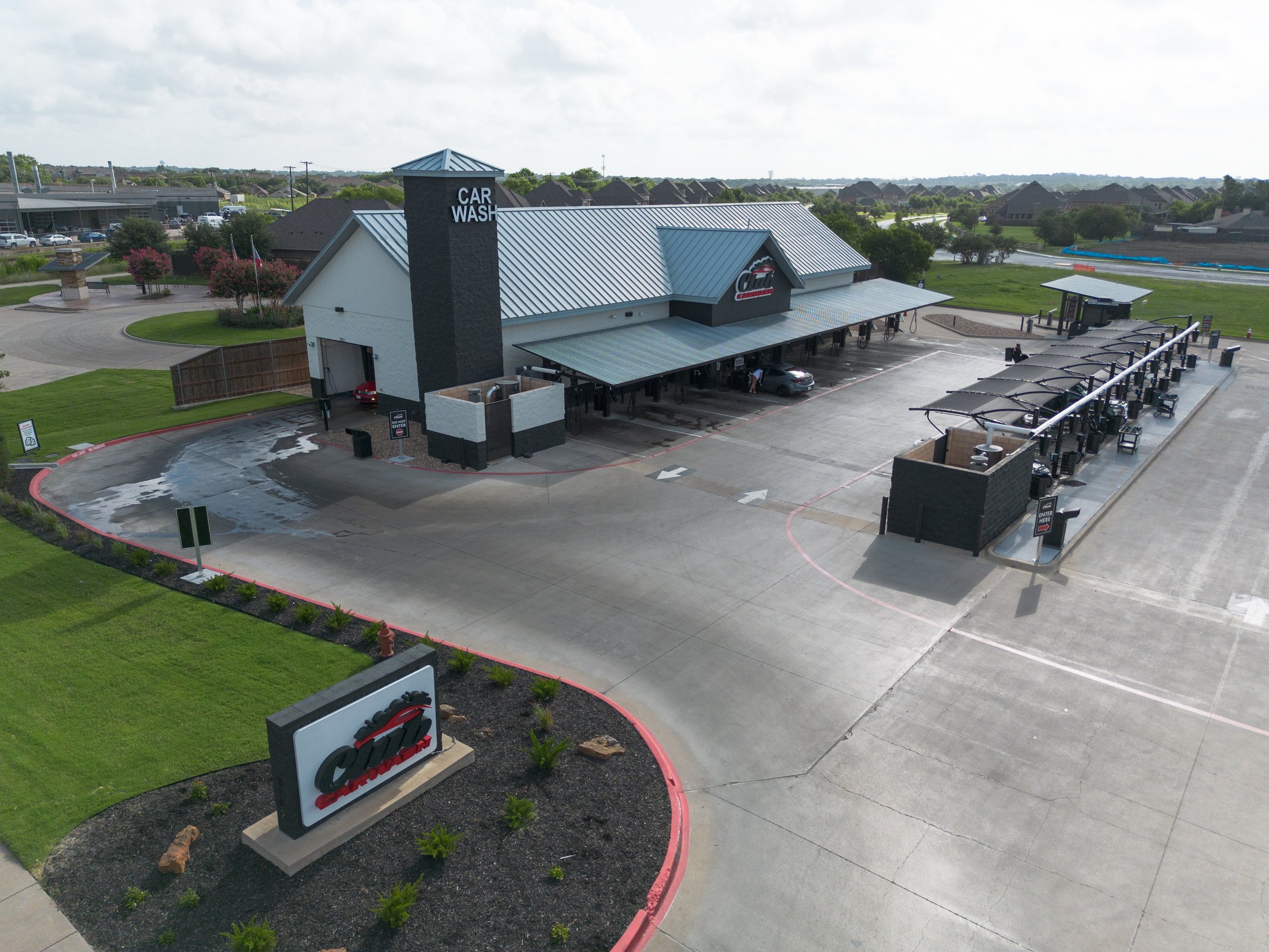 A drive-thru car wash facility with a building and multiple car wash bays, surrounded by green lawns and trees, under a cloudy sky.