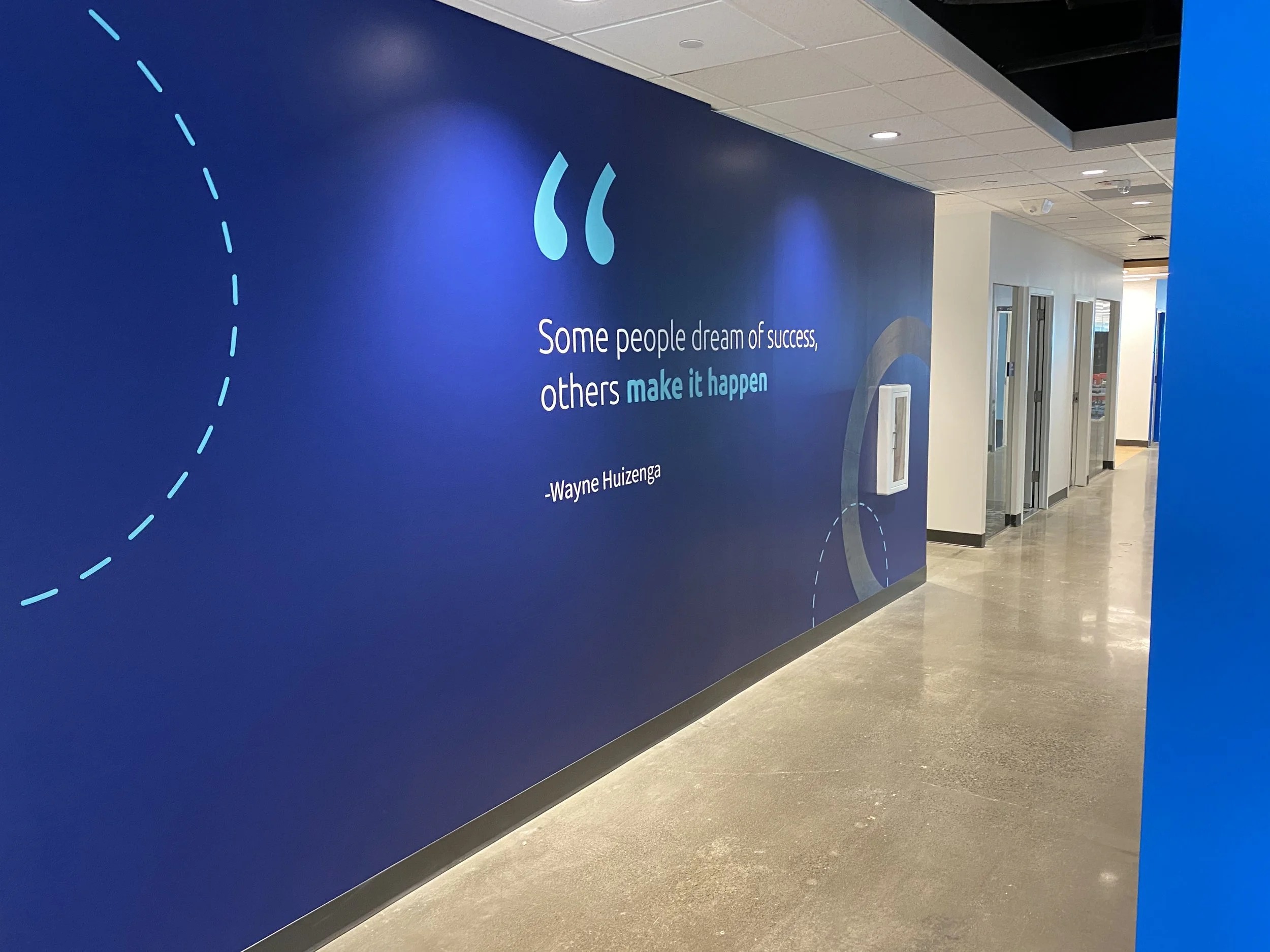 Blue wall with an inspirational quote: "Some people dream of success, others make it happen" by Wayne Huizenga, with decorative dotted lines and a fire extinguisher mounted on the wall in a corridor.