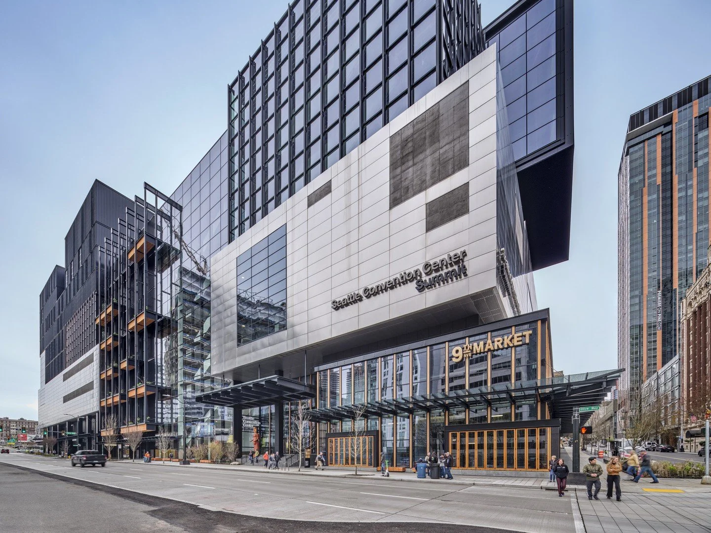 Exterior view of the Seattle Convention Center with modern glass and metal architecture, signage for the 9th Avenue Market, and pedestrians along the street in Seattle, Washington.