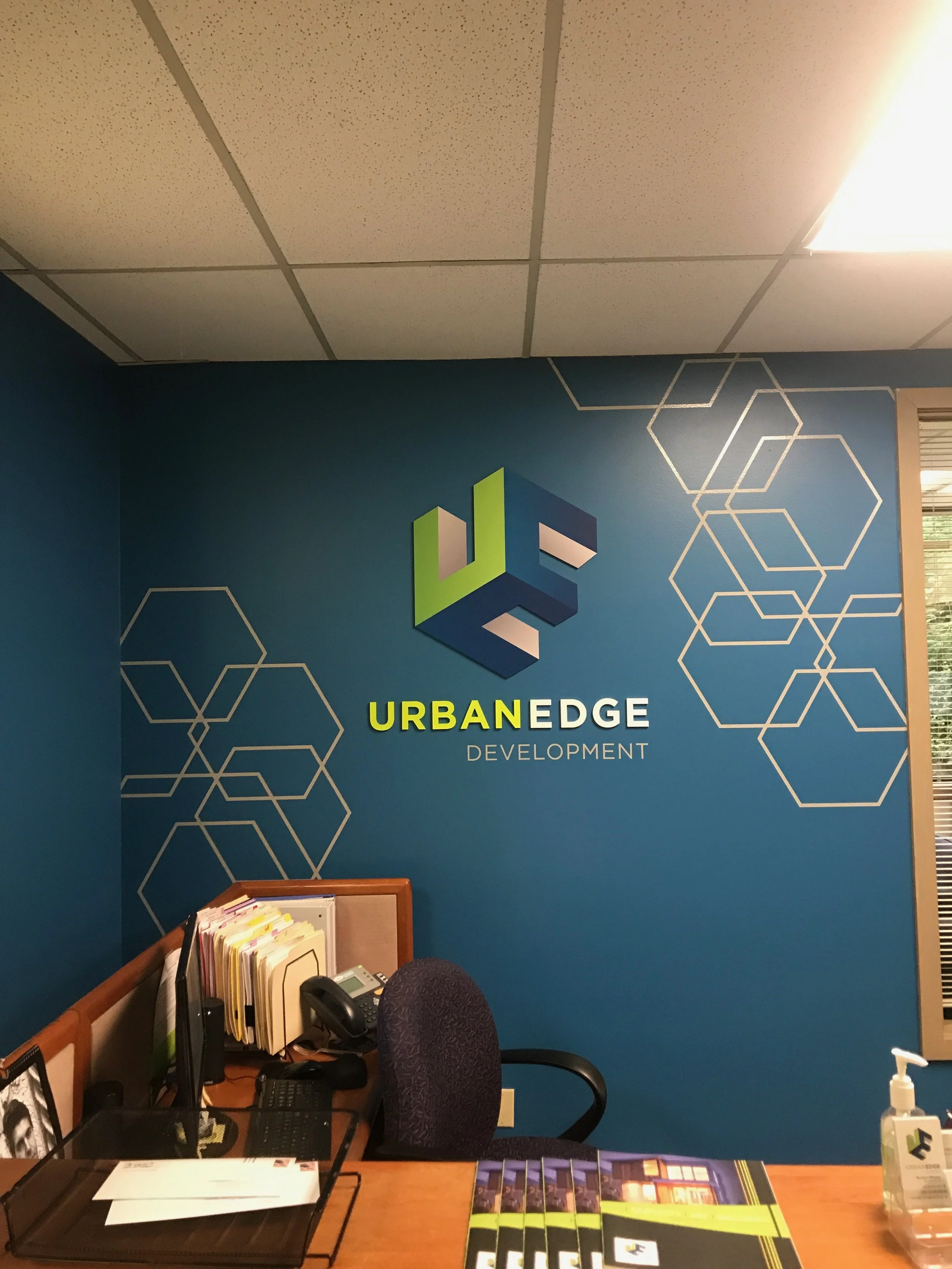 Office reception area with blue wall displaying the Urban Edge Development logo and name, with honeycomb pattern decals, a desk with papers, a computer, a telephone, an office chair, and a hand sanitizer bottle.
